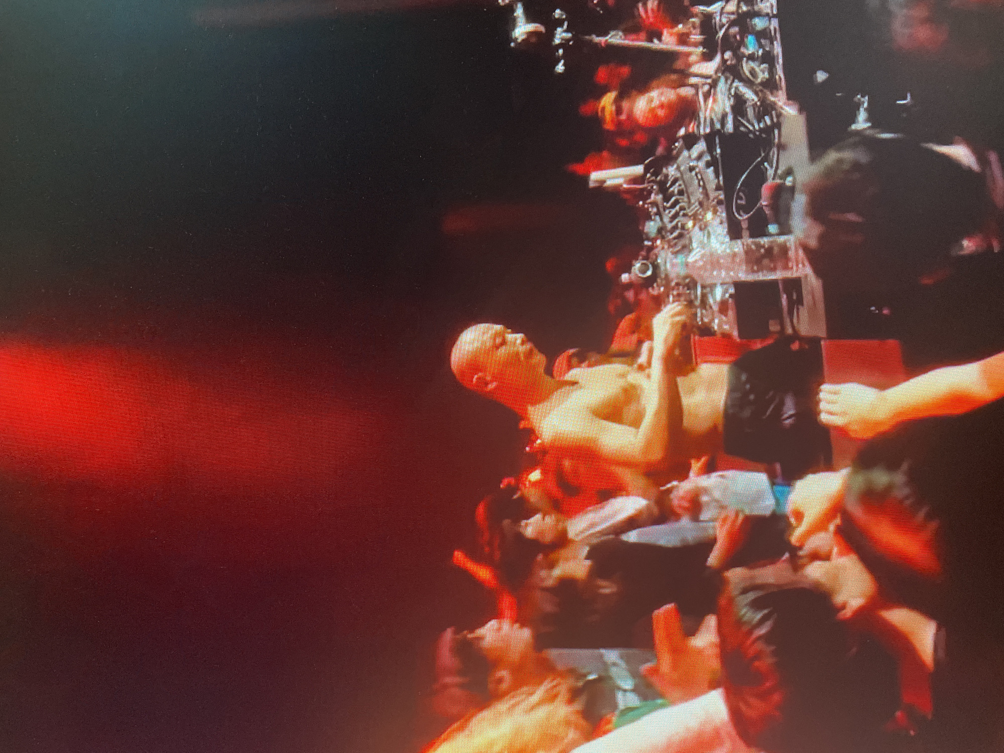 The image shows a crowded indoor venue, possibly a nightclub or concert hall, illuminated by red and white lights. In the center, a bald, shirtless man is standing behind a DJ setup, appearing to be mixing music. He is wearing dark shorts. Around him, a diverse crowd of people are visible, some with their hands raised, suggesting an energetic atmosphere. The background is dark, with additional stage lighting casting shadows and highlights. The overall impression is a lively performance or party setting. The location is in the city of Unknown, Russia.