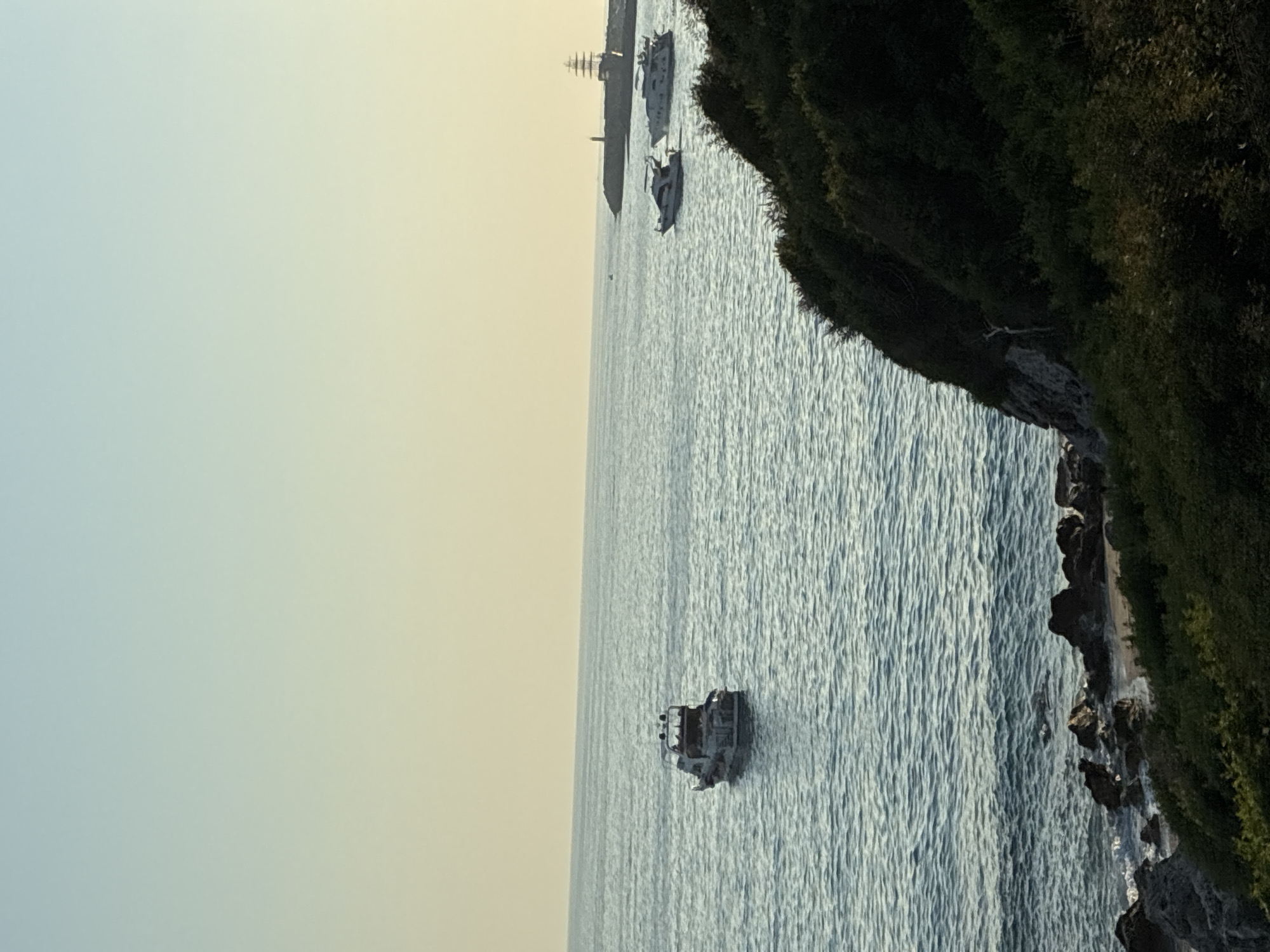 The image shows a body of water with several boats. In the foreground, there is a rocky shoreline with green vegetation. The sea is calm with gentle waves. In the mid-ground, a small white motorboat is visible. Further in the distance, two larger white motorboats are sailing. On the right side of the image, a breakwater extends into the sea, with a lighthouse and a tall, mast-like structure, possibly a historical sailing ship replica, situated on it. The sky is a pale yellow-orange, indicating it might be sunrise or sunset. The scene appears to be a coastal area. Without additional information, the specific location within Alanya, Turkey, cannot be definitively determined, but it is likely a view of the Mediterranean coast near the city. There are no people visible in the image.