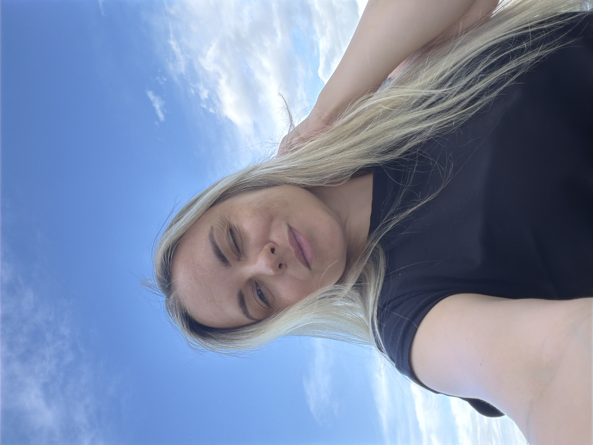 A woman with blonde hair, wearing a black shirt, is looking downward, with the sky visible in the background. The sky is blue with scattered white clouds. The woman's left eye is closed in a wink. No further details can be determined about the location in Votkinsk, Russia.