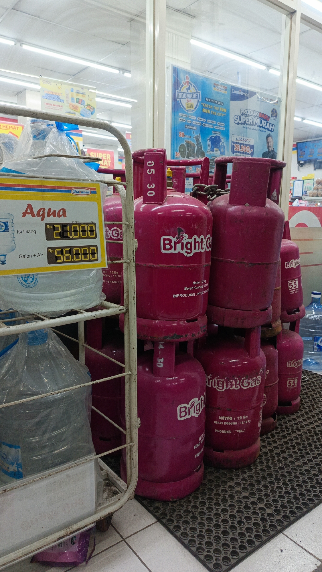 The image depicts a retail store, likely a convenience store, with a focus on a display of propane gas cylinders. The store appears to be well-lit with fluorescent lighting. In the foreground, a metal display rack holds large bottles of water, some wrapped in plastic. A yellow price tag is affixed to the rack, advertising "Aqua" with prices for refill and gallon+air. Behind the water bottles, a prominent display of bright pink propane gas cylinders is visible. These cylinders are stacked and arranged for sale. Some cylinders have labels indicating "Bright Gas." The bottom portion of the cylinders rests on a black, textured mat. In the background, shelves with various products are visible, along with signage. One sign in the background is for "Indomaret," a popular Indonesian convenience store chain, suggesting the location is likely within one of their outlets. Another sign promotes a "Fun Run" event. The overall scene conveys a typical retail environment where household goods, including cooking gas and bottled water, are sold. The image does not contain any people. The location is identified as Margajaya, Indonesia.