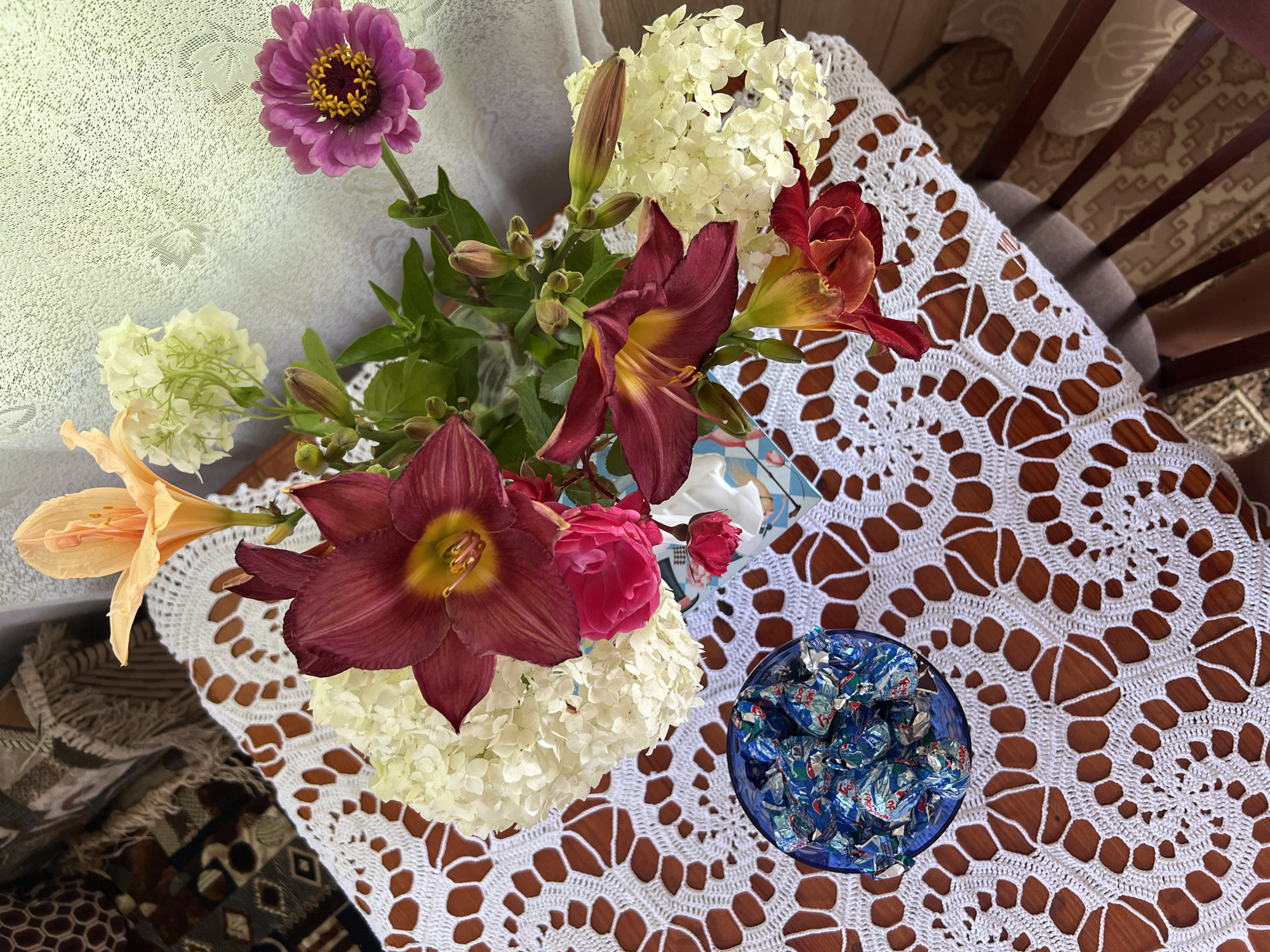 The image shows a close-up, overhead view of a wooden table adorned with a white crocheted lace tablecloth. On the tablecloth, a bouquet of flowers is arranged in a vase, featuring deep red lilies, pink roses, white hydrangeas, and an orange lily. To the left of the flowers, a small blue bowl is filled with individually wrapped candies. The background is slightly out of focus, revealing patterned textiles, possibly upholstery, and a textured wall. The scene appears to be indoors, likely a domestic setting. The image does not contain any information about the city of Lopatino, Russia.