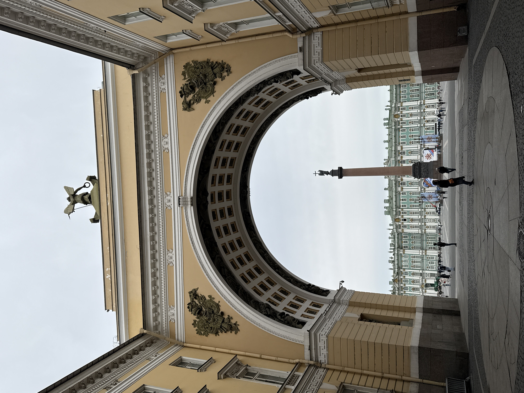 The image shows a view through the General Staff Building archway in Saint Petersburg, Russia. The Alexander Column and the Winter Palace are visible through the arch. The archway is decorated with architectural details, including moldings and sculptures. People are walking in the square beyond the archway. The ground is paved with stone. The sky is overcast.