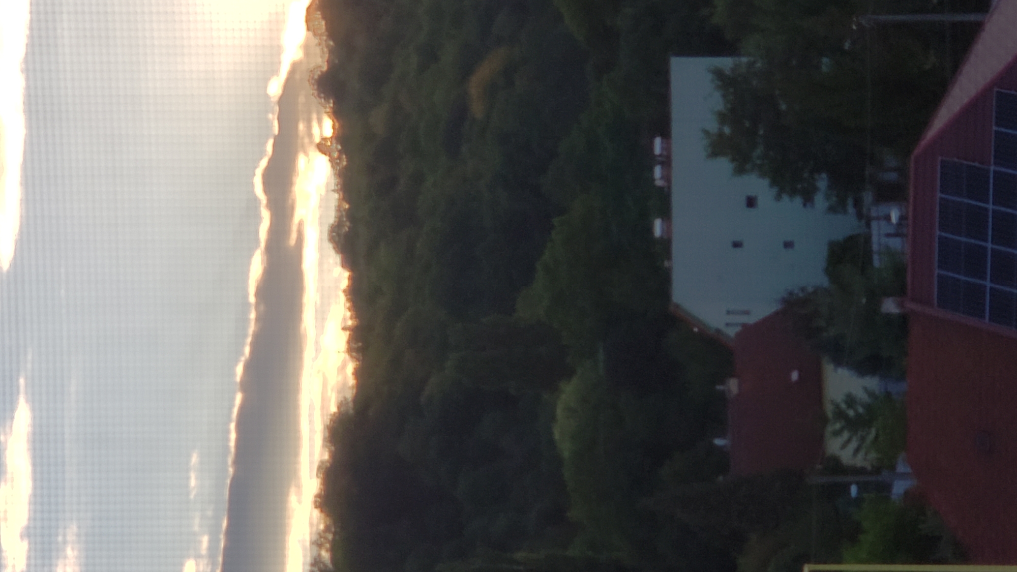 The image captures a suburban or semi-rural landscape at dusk, likely from an elevated viewpoint. The sky is a pale blue transitioning to a pale orange at the horizon, with scattered clouds. The sun appears to be setting behind a densely wooded hill that dominates the midground. The hill is covered in dark green foliage, indicating a significant amount of trees.
In the foreground, there are several buildings with red tile roofs. One building, situated to the left of center, is a light brown color with a dark red roof and appears to be a smaller, possibly residential structure. To the right of this, a larger, multi-story building with a light blue-green facade is visible. This building has several rectangular windows and appears to be an apartment complex or a larger institutional building. The roof of another building, covered in solar panels, is visible in the lower right portion of the frame.
The scene suggests a calm, residential area on the outskirts of a city, with a prominent natural landscape feature (the wooded hill) providing a backdrop. The time of day is likely late afternoon or early evening, given the soft lighting and the position of the sun. There are no people visible in the image. The image is slightly pixelated, possibly due to low resolution or being taken through a screen.