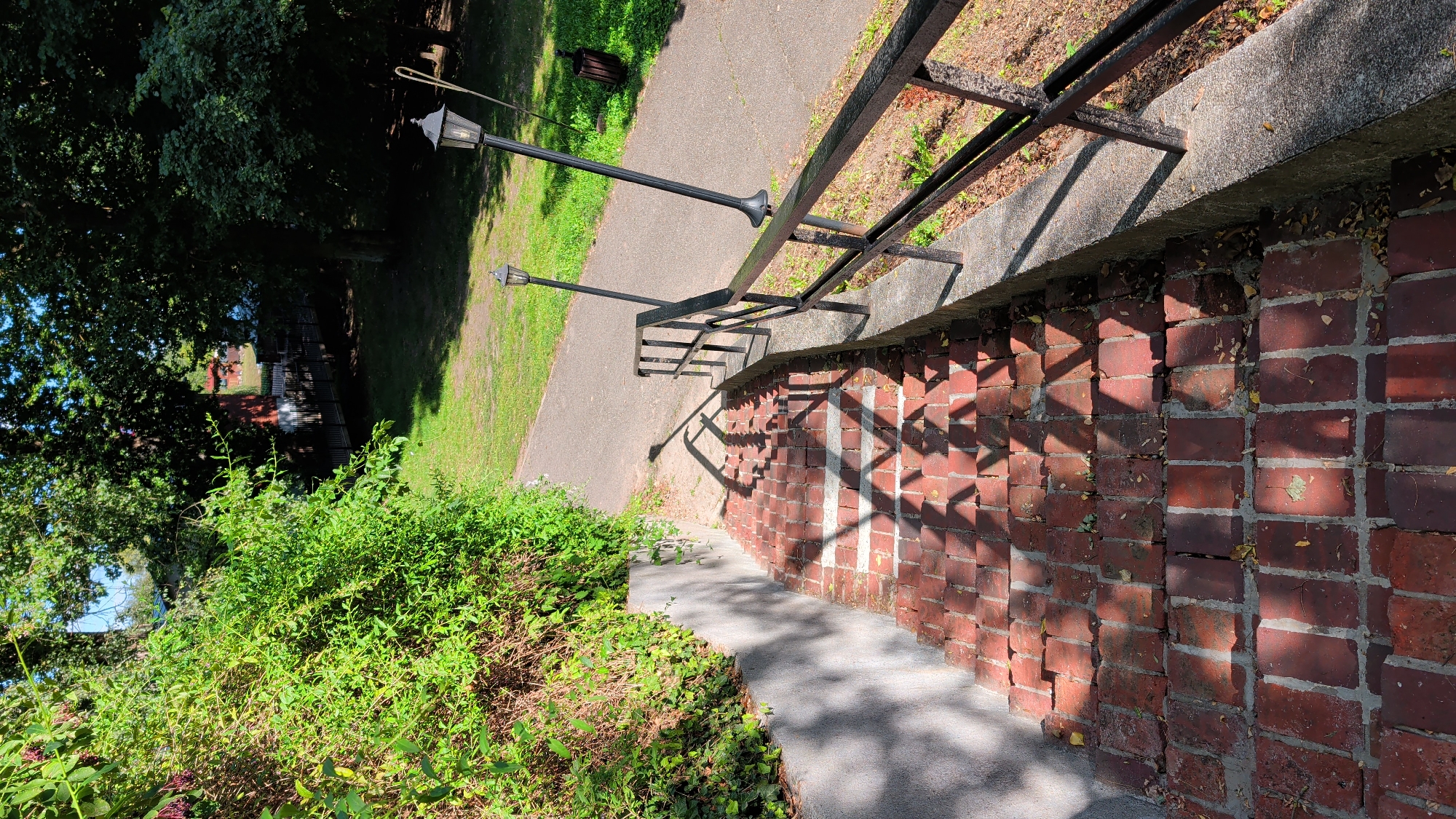 A brick staircase descends into a park area. Lush green foliage lines the left side of the staircase, with a grey concrete retaining wall. A black metal railing runs along the right side of the stairs, with two black lampposts visible further up the path. A paved pathway curves away to the right, with grass and trees in the background. The sun casts shadows across the brick steps. There are no people visible in the image. The location is not specified as Pułtusk, Poland.