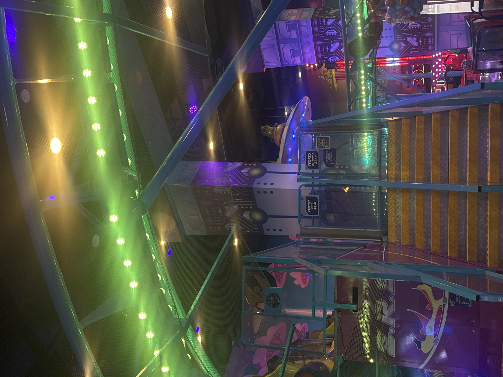 The image shows the interior of a brightly lit indoor playground. The playground structure is multi-tiered and made of metal framing with netting and panels. It features stairs with yellow and gray treads, walkways, and decorative elements with cartoonish designs. The lighting includes green LED strips, and there are signs in Hebrew and English. The setting suggests an entertainment venue for children in Tel Aviv, Israel, given the Hebrew signage and the nature of the playground. No people are visible in the frame.