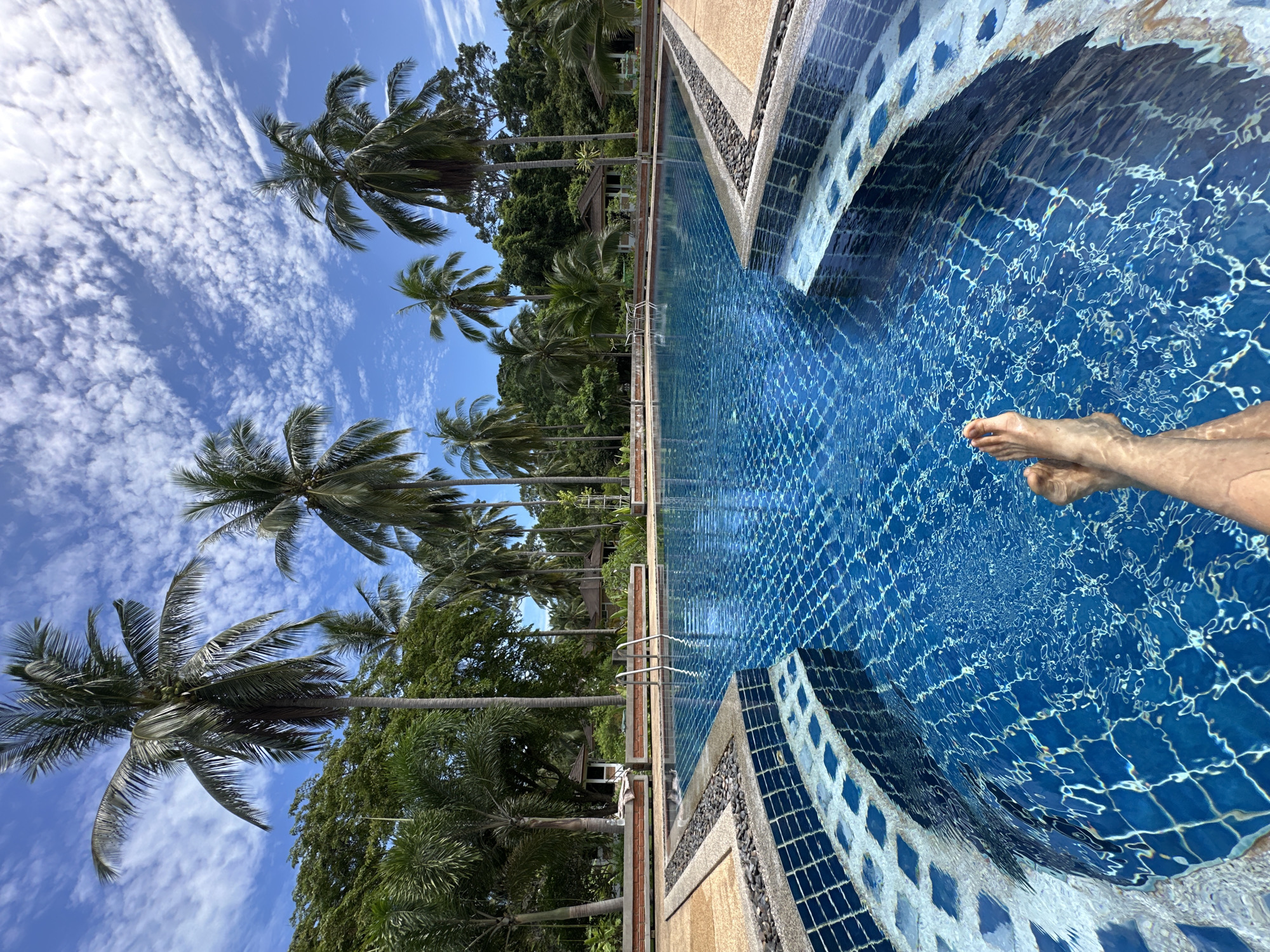 The image shows a first-person perspective of a person's legs submerged in a bright blue swimming pool, with a view of palm trees and a clear sky in the background. The pool has a tiled edge and is partially covered by a netting. Beyond the pool, lush green vegetation and several buildings with brown roofs are visible, suggesting a tropical resort or residential area in Baan Lipa Noi, Thailand. The sky is blue with scattered white clouds.