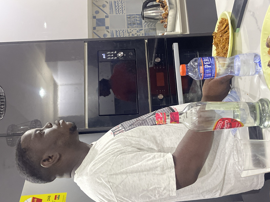 This vertical, eye-level indoor shot depicts a man in a modern kitchen, likely located in Adenta Municipality, Ghana. The subject is a Black man, seen in profile facing left, with short dark hair and a neatly trimmed beard. He is wearing a light-colored, short-sleeved t-shirt, and his gaze is directed slightly upwards.
His right arm is visible in the foreground, near two bottles. One is a clear plastic water bottle with a blue and orange label showing "PERLE." Next to it is a clear glass bottle with a red and black label, identifying the brand as "KASAPREKO" and the flavor as "RIVAL Cranyberry," with additional text "HERBA SAX." A clear plastic cup is positioned below these bottles.
The kitchen background features dark, built-in appliances, including an oven or microwave with a digital display clearly showing "10:00." Above the appliances, there is a white countertop and a decorative tiled backsplash with blue and white geometric patterns. A silver electric kettle is visible on the countertop further back. To the right, a yellow plate holds a serving of food, resembling fried plantains or a similar snack. A portion of a yellow and green energy efficiency label is visible on the far left, displaying "248" and the word "GHANA," which helps confirm the location. The scene suggests a casual moment in a domestic kitchen, possibly during meal preparation or a social gathering.