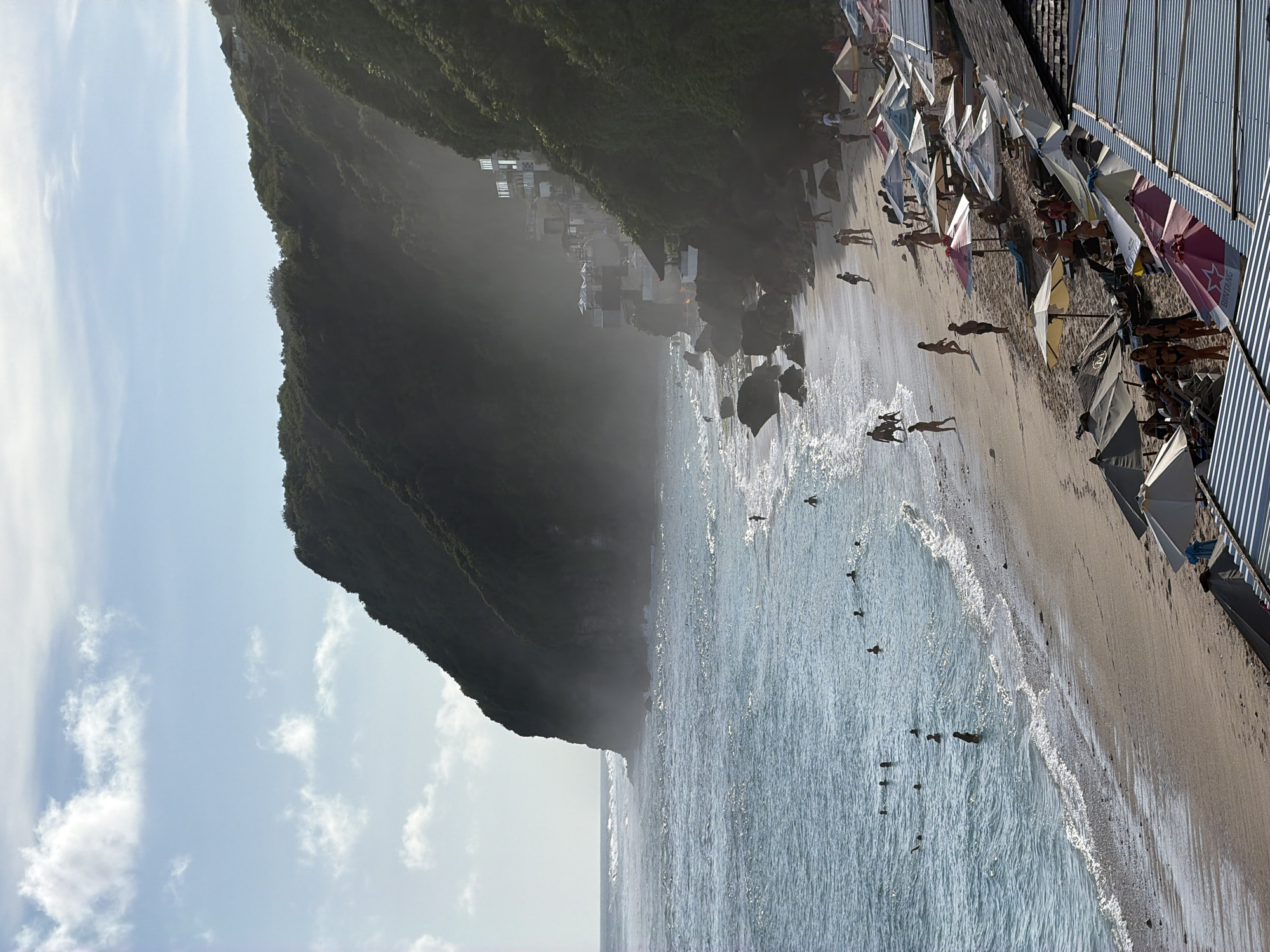 This photograph shows a beach in Kuta Selatan, Indonesia. A lush green cliff rises sharply from the left of the frame. Houses and buildings dot the cliffside. The beach extends along the right side, populated with people and beach umbrellas. Waves break on the shore. Swimmers are visible in the water. The sky is light blue with scattered clouds.