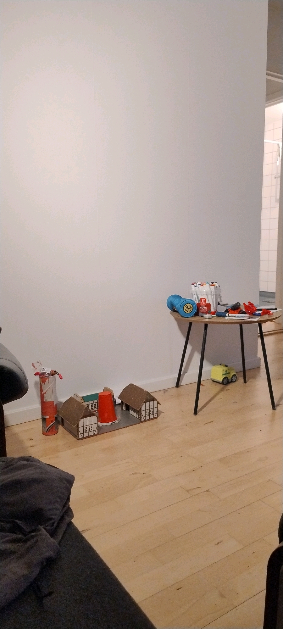 The image shows a living room in Ishøj, Denmark, with a focus on toys and a small table. The room has light-colored walls and a light wooden floor. In the foreground, a dark gray sofa is visible on the left. Further back, a diorama of small houses with brown roofs and white-trimmed walls sits on the floor. To the left of the diorama is an orange cylindrical object with red and white markings, possibly a toy fire extinguisher, with some small toys inside. To the right of the diorama, a red plastic cup is placed in the center. In the background, a small, round table with black legs holds various items, including a blue toy, a stack of markers, and some red toy accessories. A small yellow toy vehicle is on the floor near the table. The overall impression is a child's play area in a residential setting.