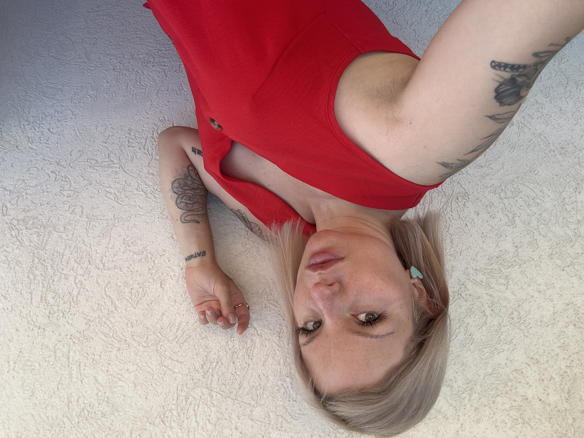 The image shows a woman lying down, with her head tilted back. She is wearing a red dress and has blonde hair. Her left arm is visible, with tattoos on it and the word "GATHER" tattooed on her forearm. She is wearing a ring on her left hand and a heart-shaped earring in her right ear. The background is a textured white surface. The lighting suggests it is daytime, possibly outdoors given the textured surface which could be a wall or ground. There is no clear indication of weather. The setting is not definitively identifiable as Budva, Montenegro, based solely on the visual information provided in the image. The woman's expression is neutral.