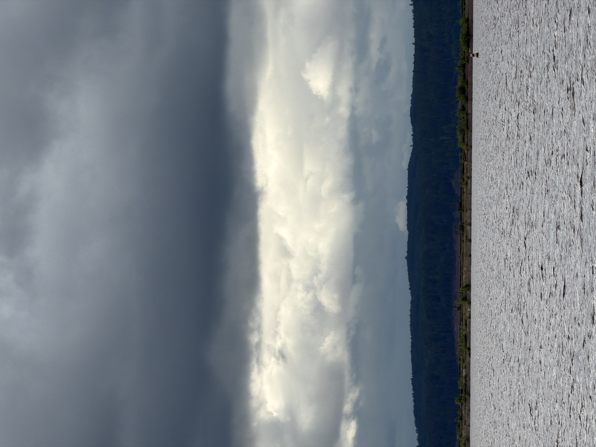 The image depicts a vast outdoor landscape featuring a body of water, a distant forested shoreline, and a heavily clouded sky, captured in the vicinity of Chaikovsky, Russia. The sky, occupying approximately two-thirds of the frame, is stratified. The uppermost section features a dense, dark grey cloud layer. Below this is a pronounced horizontal band of brighter, off-white clouds, indicating a break or reflection of light. Beneath the bright band, another layer of darker, more diffuse grey clouds transitions towards the horizon.
The horizon line reveals a distant, dark green, undulating landmass densely covered with trees, characteristic of a forested hill or mountain range. Below this, closer to the water's edge, a flatter, lighter-colored strip of ground is visible, possibly a bank or embankment, with sparse green vegetation. Vertical pole-like structures are faintly discernible extending from this strip towards the lower cloud layer.
The lower third of the image is dominated by a wide expanse of water, appearing grey with numerous small, uniform ripples on its surface, indicative of gentle movement and reflecting the overcast light. In the lower right quadrant of the water, a single navigational buoy is present. It is cylindrical, displaying distinct horizontal red and white stripes, topped with a small, dark finial. No people, human activity, or direct interactions are visible within the scene.