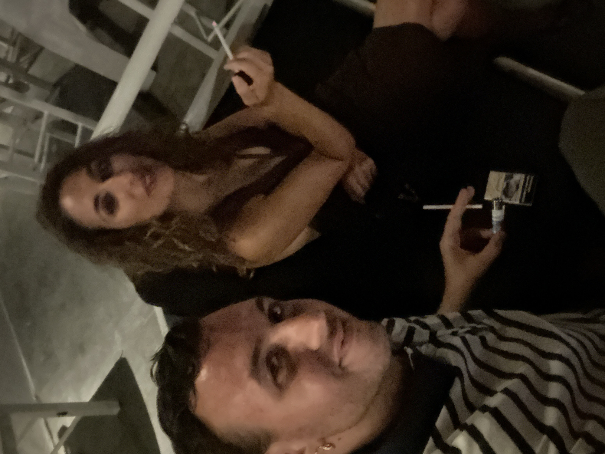 An outdoor, nighttime portrait shows a man and a woman seated in what appears to be a lounge area with white chairs and tables. The man is in the foreground, to the left, wearing a striped shirt and looking at the camera with a slight smile. He has dark hair and a gold hoop earring in his left ear. The woman is seated behind and to the right of the man, looking slightly away from the camera with her right hand holding a lit cigarette. She has long, curly brown hair and is wearing a dark dress. A pack of cigarettes and a lighter are visible on the dark surface in front of them. The scene suggests a casual social gathering in Mykonos, Greece, possibly at a bar or restaurant given the seating arrangement and ambiance.