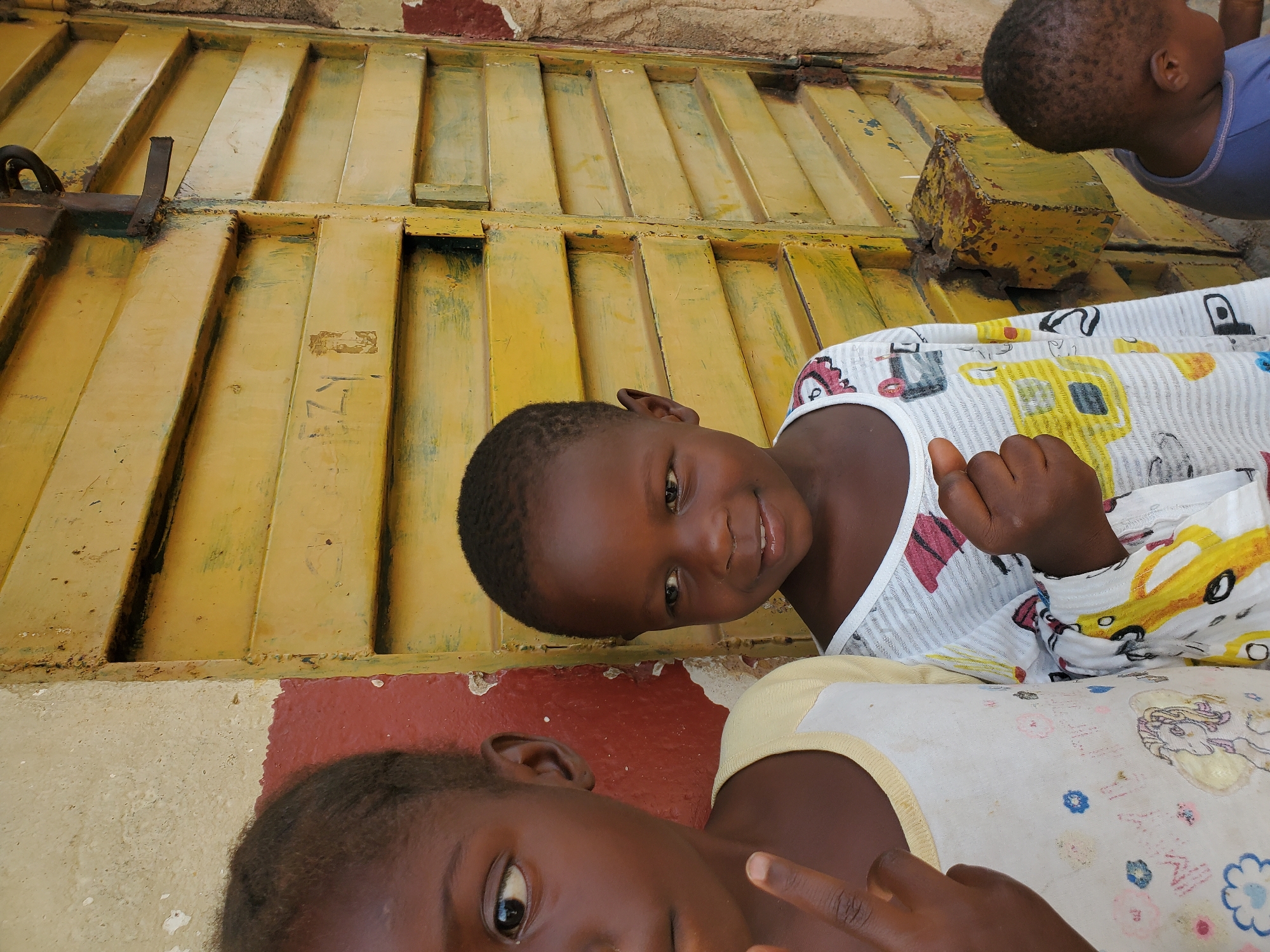 A medium shot captures three young children standing in front of a weathered, bright yellow corrugated metal door, likely in an urban setting. The door is in a state of disrepair, with chipped paint and visible rust. To the left of the door, a beige and red textured wall stands, also showing signs of wear.
The child in the foreground on the left, a girl, is partially visible, with dark skin and hair. She is looking to her left and her left hand is raised, with two fingers extended in a peace sign. She wears a white long-sleeved shirt with a yellow collar adorned with colorful cartoon prints.
To her right, another child, also a girl, is more fully visible. She has dark skin and hair and is looking towards her right with a slight smile. She is wearing a white long-sleeved shirt with a more complex pattern of colorful cartoon elements. Her right hand is clenched in a fist.
A third child, a boy, is visible in the background on the far right, his head and shoulders appearing in the frame. He has dark skin and hair and is looking downwards. He wears a blue t-shirt.
The overall impression is one of a snapshot of everyday life, possibly in a neighborhood of Jos, Nigeria, given the context. The environment appears to be a street or an exterior wall of a building, with the subjects positioned in a casual manner.
