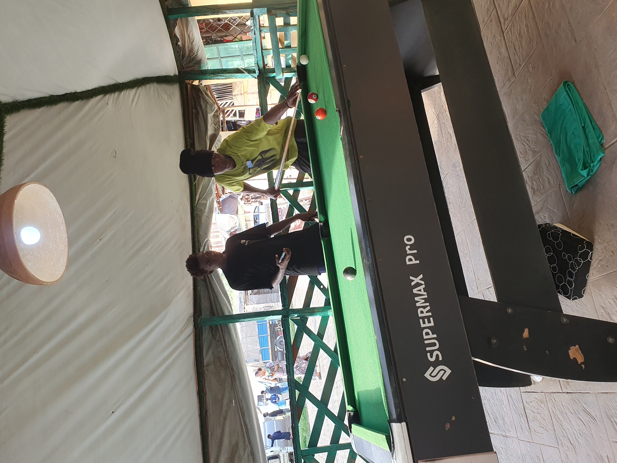 A young man wearing a lime green t-shirt and a black beanie is playing pool at an outdoor pool table. He is in the process of taking a shot, cue stick in hand, aimed at a red and white striped ball. Another young man in a black t-shirt stands to his left, observing the game. The pool table, labeled "SUPERMAX Pro," has a green felt surface and several balls are scattered across it, including a white cue ball and a solid black ball. The setting appears to be a covered outdoor area, possibly a bar or recreational space, with a white tent-like ceiling overhead. In the background, there are glimpses of other people and vehicles, suggesting a public area within the city of Lokoja, Nigeria. The ground is paved with textured tiles, and a folded green cloth is visible beneath the pool table.