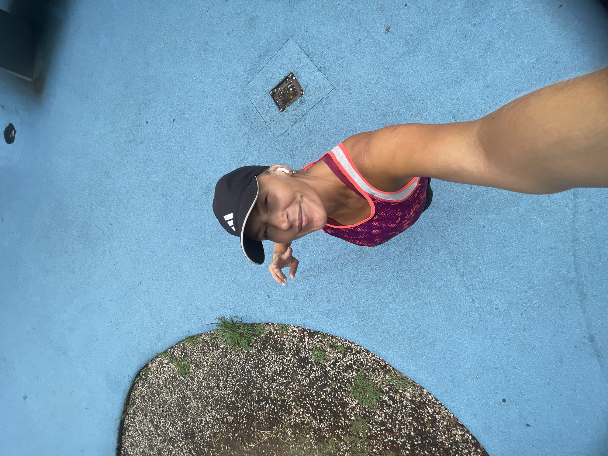 A woman with brown hair, wearing a black baseball cap with an Adidas logo and a purple tank top, stands on a blue paved surface. She is holding a phone in her right hand, taking a selfie. In the background, there is a patch of gravel with some sparse green vegetation. A square metal drain cover is visible on the blue surface to her right. The scene appears to be outdoors, possibly in a park or recreational area in Sochi, Russia, judging by the texture of the paving and the presence of the drain cover.