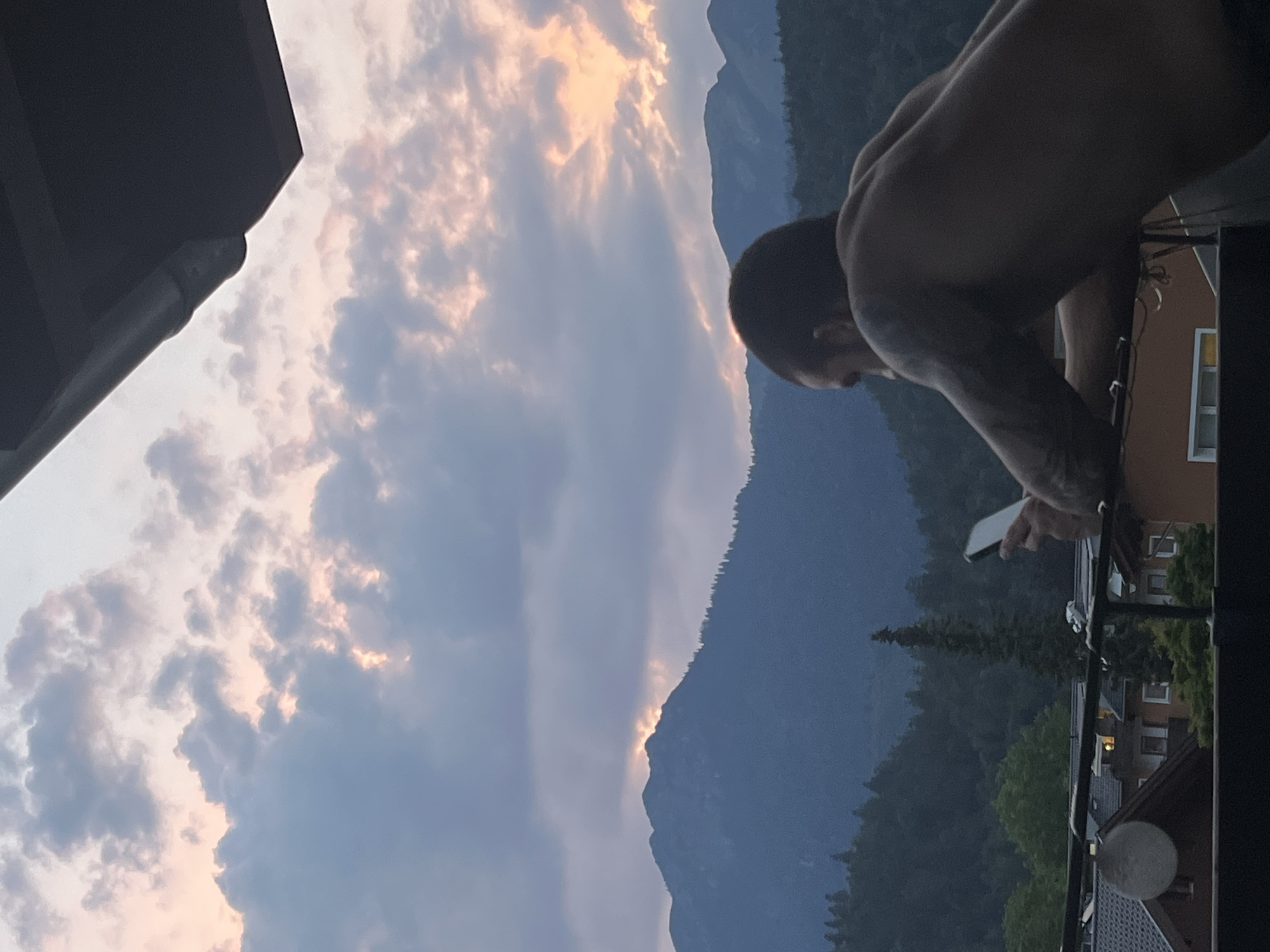 The image depicts a person on a balcony in Garmisch-Partenkirchen, Germany, holding a smartphone. The scene is set against a backdrop of a dramatic sky with sunlit clouds and silhouetted mountains. The person is shirtless, with visible tattoos on their arm, and is looking down at their phone. The time of day appears to be either dawn or dusk, given the soft, warm light illuminating the clouds and the darkening sky. The surrounding landscape includes dense evergreen trees and buildings, suggesting a residential area nestled within a mountainous region. The overall mood is one of quiet contemplation or engagement with technology amidst a beautiful natural setting. No visible text is present in the image.