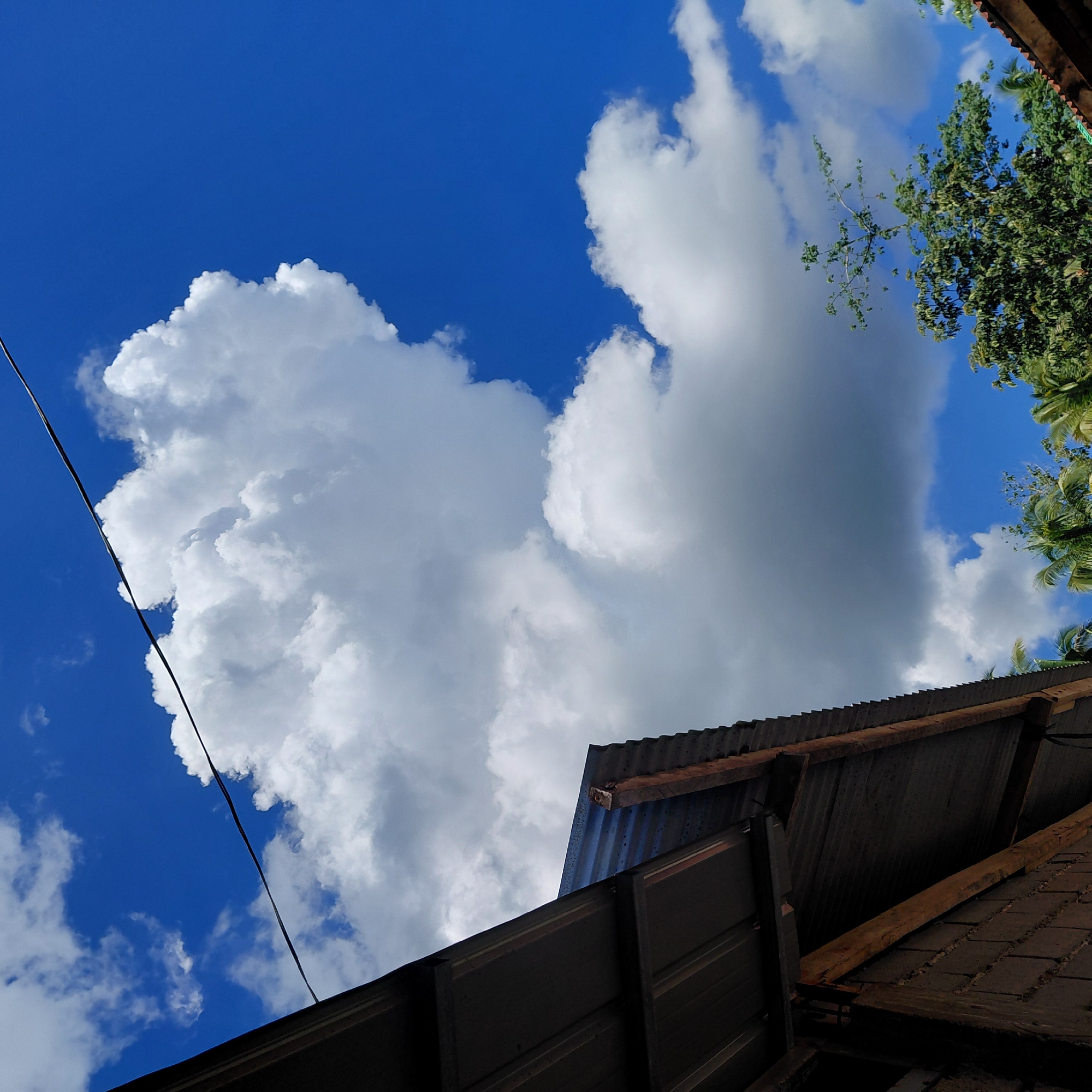 A wide-angle, low-angle shot captures a bright blue sky with dramatic cumulus clouds and a portion of a corrugated metal roofed structure in the foreground. The structure, appearing to be a building or shelter, is constructed with brick and wood, with a dark, weathered corrugated metal roof. A dark electrical wire stretches diagonally across the upper portion of the frame. In the lower right quadrant, the tops of lush green trees, including palm trees, are visible against the sky. The scene is set outdoors, likely in Samal, Philippines, given the tropical foliage and building materials. The lighting suggests it is daytime, with the sun illuminating the clouds and sky. There are no people visible in the image.