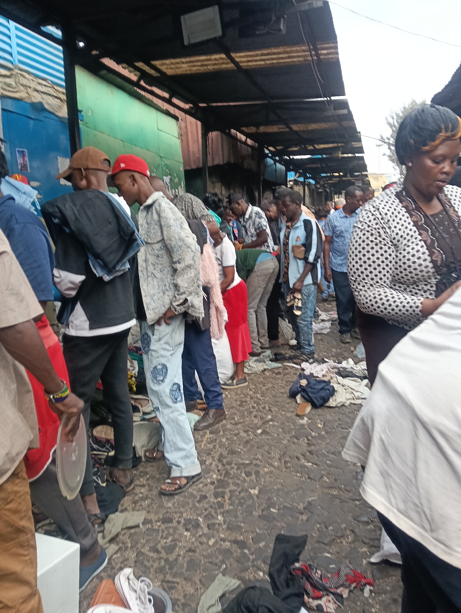This is a low-angle, slightly blurry, outdoor photograph taken in what appears to be a bustling marketplace in Nairobi, Kenya. The scene depicts a crowded area under a corrugated metal awning, suggesting a covered market or street vendors. People are seen in various states of activity, many looking at or interacting with goods laid out on the ground or on low surfaces.
In the foreground, a person's legs are visible, wearing grey pants and blue sandals. To their left, another person wears a red t-shirt and grey pants, holding a clear plastic plate. Further back, a young person in a light-colored, patterned jacket and light blue jeans stands looking down, with a red baseball cap. Beside them, a person in a black jacket and dark pants is also visible.
The ground is rough, possibly a dirt or unpaved road, with numerous items of clothing and shoes scattered around, indicating a second-hand clothing market. The background shows a mix of blue and green structures, possibly stalls or walls, and more people lining the market. The overall atmosphere is one of lively commerce and dense human activity. The lighting suggests it is daytime, with diffuse light.