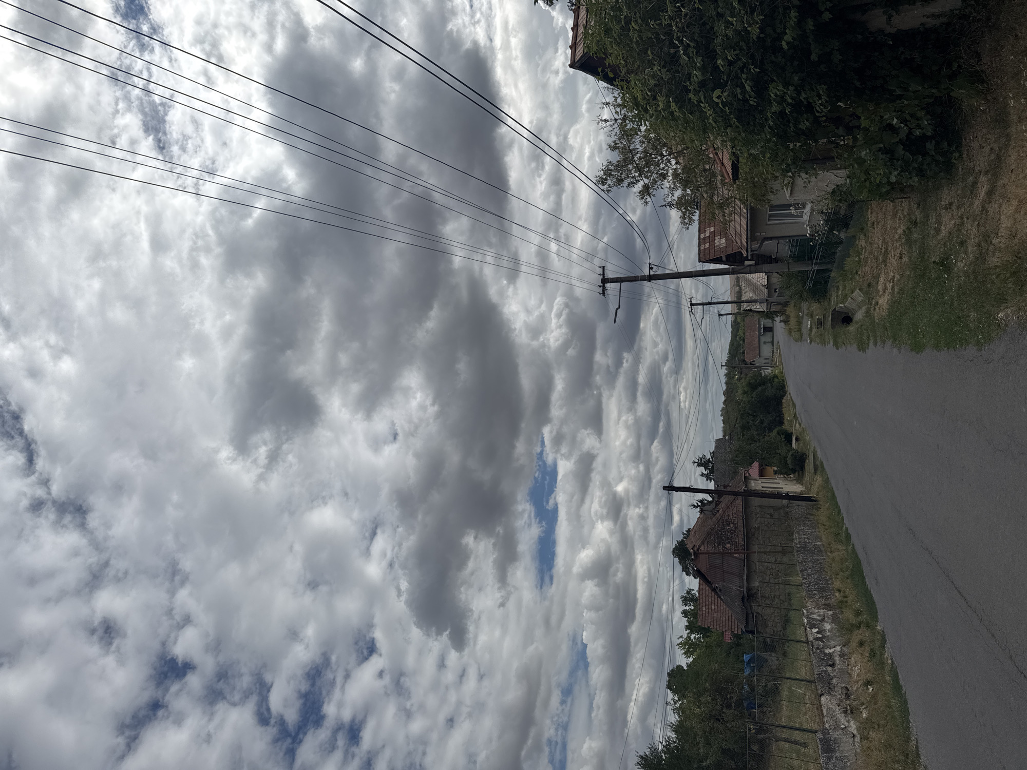 The image depicts a rural road in Hontianske Moravce, Slovakia, under a cloudy sky. The road is paved and appears to be a residential street, lined with a few houses and vegetation. On the left side of the road, there's a stone wall and a fence in front of a house with a weathered roof. On the right side, more houses are visible, some with overgrown bushes and trees. Overhead, numerous power lines crisscross the sky, supported by utility poles. The overall atmosphere is overcast and somewhat somber.
