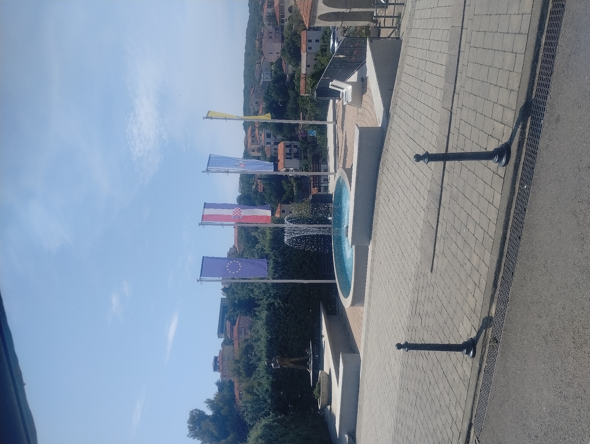 This image depicts an outdoor scene with a fountain and flags in the foreground, set against a backdrop of a town and rolling hills. The fountain is circular and appears to be made of light-colored stone, with water actively cascading from its center. In front of the fountain, a row of flags is visible, each mounted on a pole. The flags are vertical and display various colors and symbols. From left to right, the visible flags appear to be the European Union flag, followed by the Croatian flag, then a blue flag with a coat of arms, and a yellow flag. To the left of the fountain, there is a statue of a person standing on a pedestal. The area around the fountain and statue is paved with light-colored rectangular stones, forming a walkway. Two dark-colored bollards are positioned on the paved area. Beyond the paved area, there is a railing overlooking a town with buildings of various colors, predominantly red-tiled roofs. The town is situated on a hillside, with lush green trees and vegetation interspersed among the buildings. The sky above is clear and blue with a few scattered white clouds. The scene suggests a public space in a Mediterranean or coastal region. Based on the flags, specifically the Croatian flag, and the style of architecture, the location is likely in Croatia, specifically in the municipality of Općina Vrbnik.