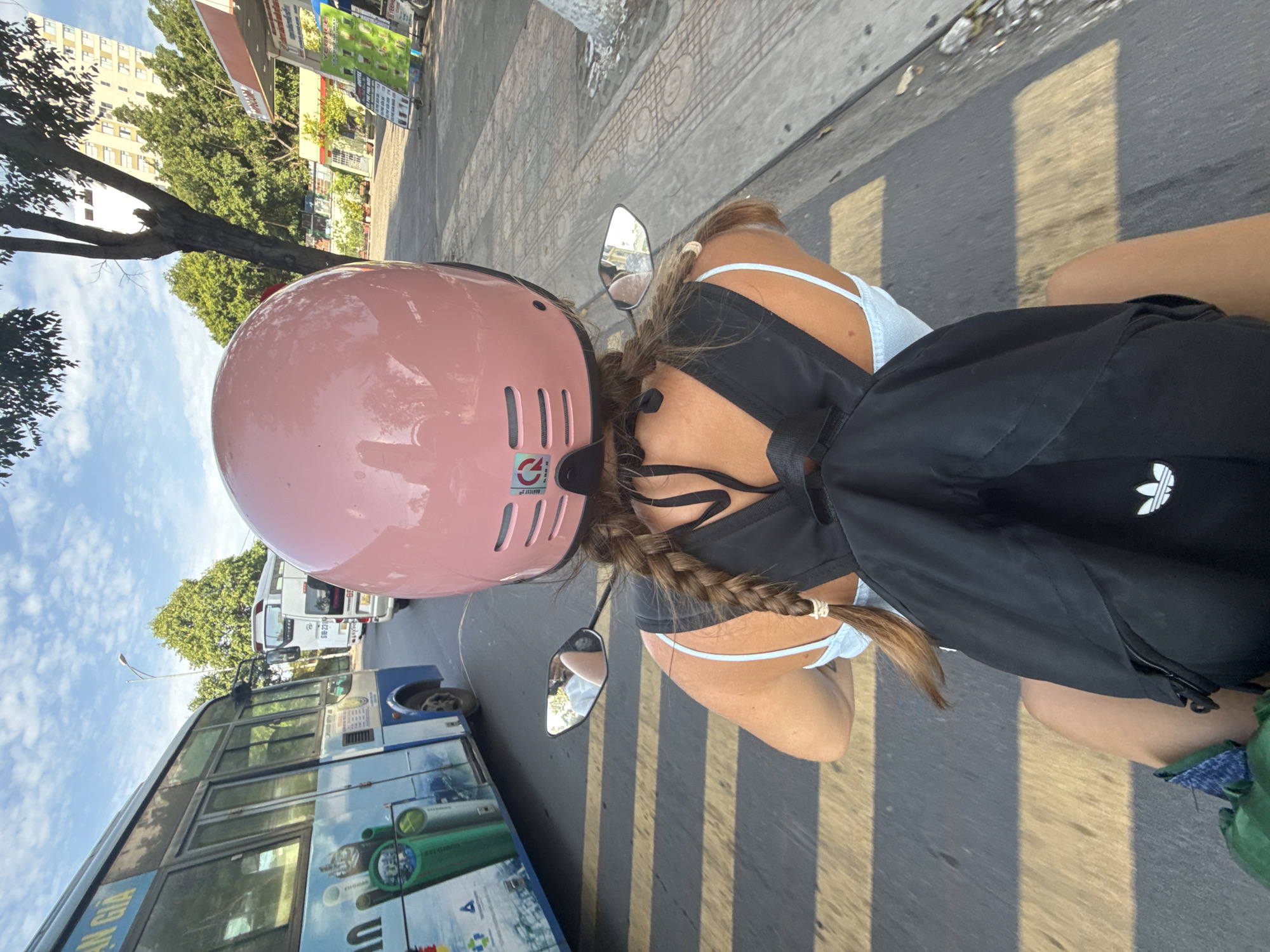 The image shows a person from behind riding a motorbike in Nha Trang, Vietnam. The rider is wearing a pink helmet, a light blue top, and a black backpack with an Adidas logo. Their hair is in a braid. To the left, a blue bus is visible, and further down the road, a gas station with signs can be seen. The scene appears to be a street in the city, with a clear sky and trees lining the road. The road has yellow and grey striped markings.
