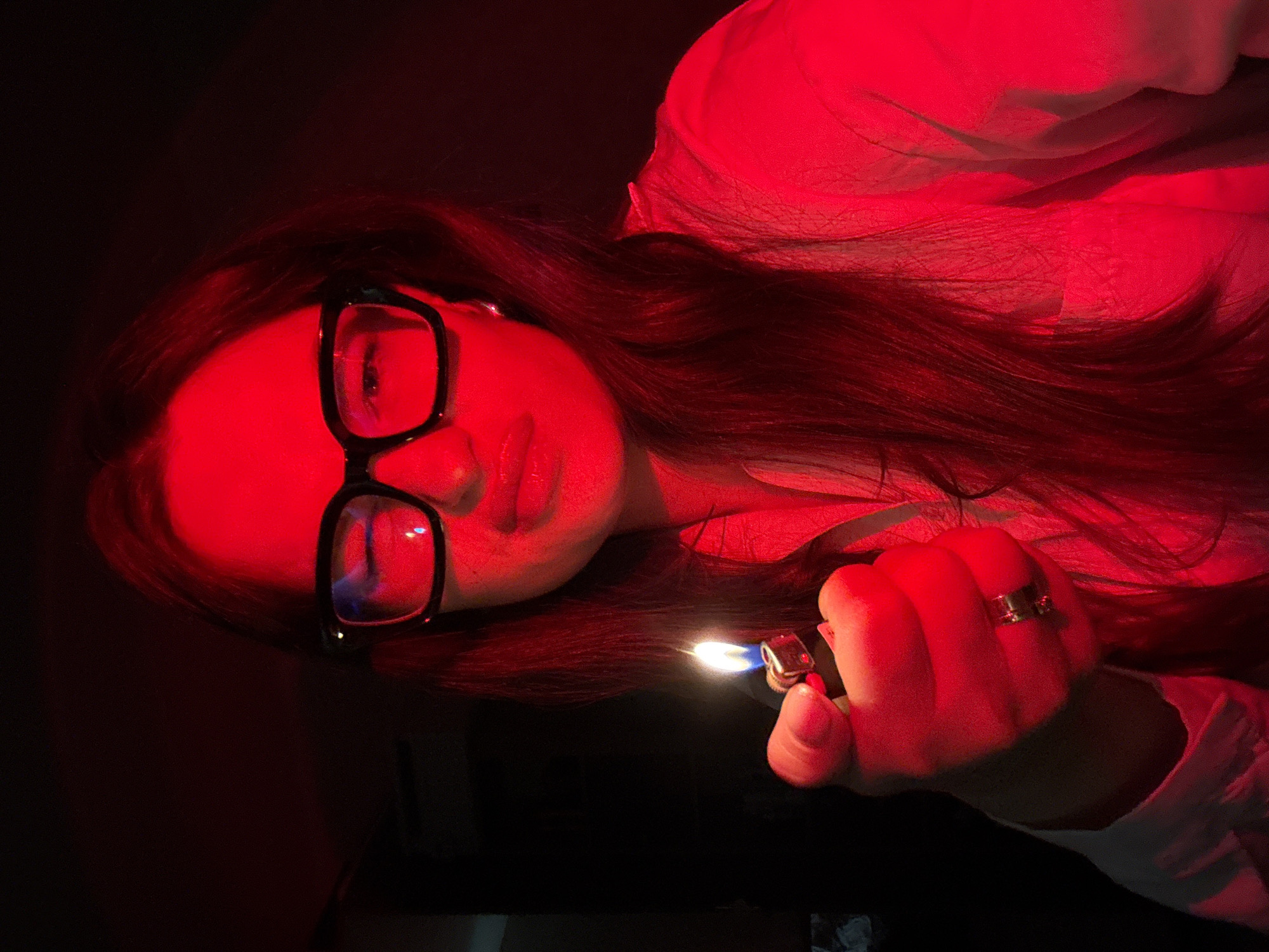 A woman with long, dark hair and black-rimmed glasses is holding a lit lighter in her right hand. She is wearing a light-colored garment, possibly a shirt or pajamas, which is illuminated by a strong red light that casts a pervasive red hue over the entire scene. Her lips are slightly parted, and she is looking towards the camera. The background is dark and indistinct. There are no discernable urban elements that would indicate a specific location within Chaikovsky, Russia; the scene appears to be an indoor setting.