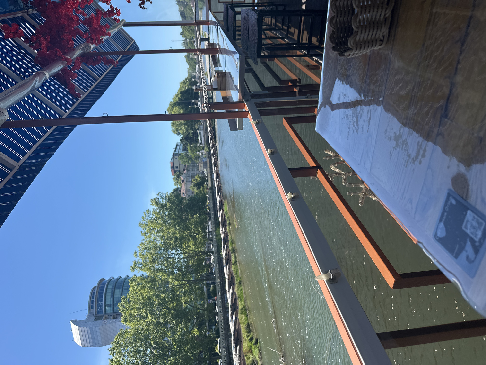 The image captures a view from a riverside restaurant in Sochi, Russia. In the foreground, a table covered with plastic wrap and a woven basket is visible. Beyond the table, a railing separates the dining area from the river. The river itself is green and calm, with a long, narrow bridge spanning its width in the mid-ground. On the opposite bank of the river, lush green trees line the waterfront, behind which are buildings, including a distinctive modern high-rise with a curved facade. The sky is a clear, bright blue, indicating a sunny day. The restaurant appears to have an outdoor seating area with a blue and white striped awning providing shade. Red artificial flowers are draped from the awning, adding a decorative touch. The scene suggests a pleasant, outdoor dining experience by the water.