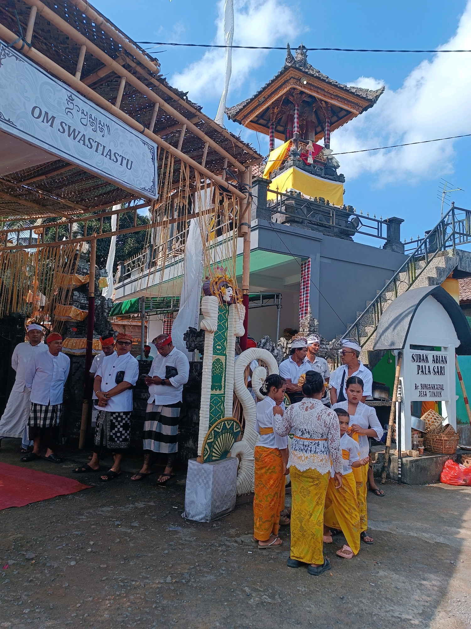 The image depicts an outdoor scene in the city of Tabanan, Indonesia, likely during a cultural or religious event. A group of people, predominantly men dressed in traditional attire including white shirts and patterned sarongs, stand near a structure adorned with bamboo and thatch roofing. Some men wear head coverings. A prominent sign with Balinese script and the text "OM SWASTIASTU" is visible.
To the right of the main group, a multi-tiered pagoda-like structure with intricate carvings and adorned with yellow and red fabric stands against a bright blue sky with scattered clouds. Further to the right, a sign that reads "SUBAK ABIAN PALA SARI" and "BR. BUNGANKAPAL DESA TUNJUK" is mounted on a white structure. Several children, mostly girls in white tops and yellow sarongs, are also present. One child is looking directly at the camera. The ground is a mixture of dirt and gravel, with a red rug laid out on the left side of the image. Various decorative elements, including woven baskets and what appears to be a figure adorned with fabric, are scattered throughout the scene. The overall atmosphere suggests a community gathering or ceremony.