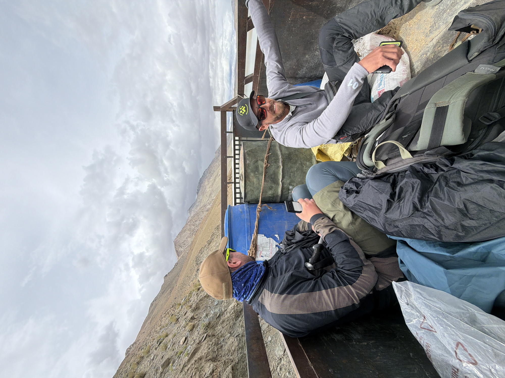 Two men are sitting in the back of a truck in a mountainous, remote area, likely near Karzok, India. The man on the left, wearing a tan baseball cap and a blue patterned neck gaiter, is looking down at his phone. He has on a black jacket with brown stripes and olive green pants. A walking stick is visible between his legs. To his right is a large blue barrel secured with rope.
The man on the right, wearing a black baseball cap with a yellow logo and sunglasses, is smiling and extending his right arm. He is wearing a grey long-sleeved shirt and black pants. He is also holding a phone in his right hand. Several backpacks and duffel bags are piled around the men in the truck bed.
The background shows a barren, rocky mountainside under a cloudy sky. The overall impression is a utilitarian mode of transport in a rugged, natural environment.