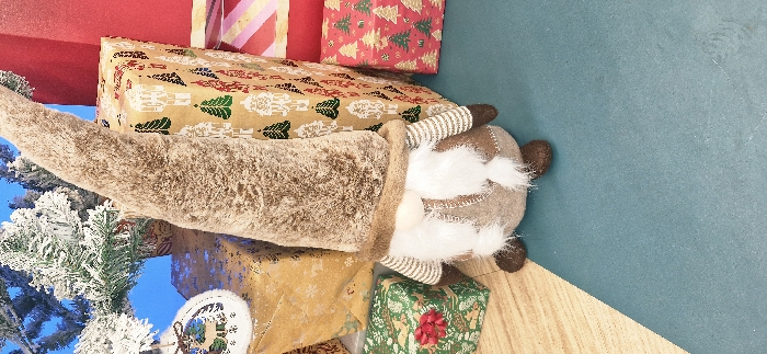 The scene depicts an indoor festive display located in Vendin-le-Vieil, France. A plush gnome figure is positioned centrally, featuring a tall, pointed, shaggy brown hat that covers its eyes, a small light-colored nose, and a prominent white beard. Its body is constructed from light brown fabric with dark stitching details, and it has striped arms with dark brown hands and rounded dark brown feet. The gnome stands on a teal-colored surface, which transitions to a light wood-grain floor on the left side of the frame. Surrounding the gnome are several wrapped gift boxes: a large vertical box wrapped in brown paper with white nutcracker and green Christmas tree patterns is to its right, alongside a smaller red box with green tree patterns. To the gnome's left, a gold-colored box with snowflake patterns is visible, and a green box with a small red bow rests further left. In the upper left corner, an artificial Christmas tree with snow-flocked branches and a white ornamental cutout of a deer hangs against a plain blue background. All subjects are inanimate and arranged in a static configuration. No human figures are present.