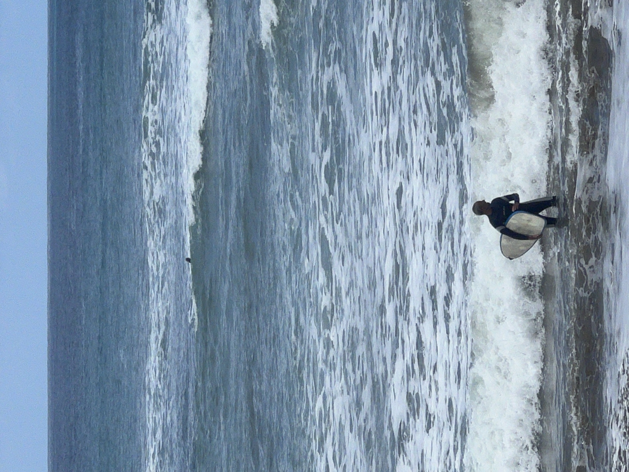 The image captures an ocean scene at a beach, likely in Las Palmas De Gran Canaria, Spain, dominated by blue water and breaking waves under a light sky. The central subject is a person, likely a bodyboarder, situated in the right half of the frame. This individual is wearing a dark, full-body wetsuit and holds a white and light blue bodyboard against their side, standing in the turbulent, foamy water where a wave is crashing.
The ocean fills most of the visual space, displaying various textures from the movement of water. On the right, close to the person, a significant amount of white foam indicates a powerful breaking wave. Further out to the left, the water appears calmer but still features ripples and patterns from current and smaller swells. A small, dark, indistinct object or person is faintly visible far out in the ocean on the mid-left side.
The sky, occupying the top portion of the image, is a soft, uniform light blue or grey, suggesting an overcast day or diffused daylight. There are no harsh shadows present, confirming it is daytime. The scene conveys the activity of waiting for or entering/exiting the waves during a surfing or bodyboarding session. No visible text is present in the image.
