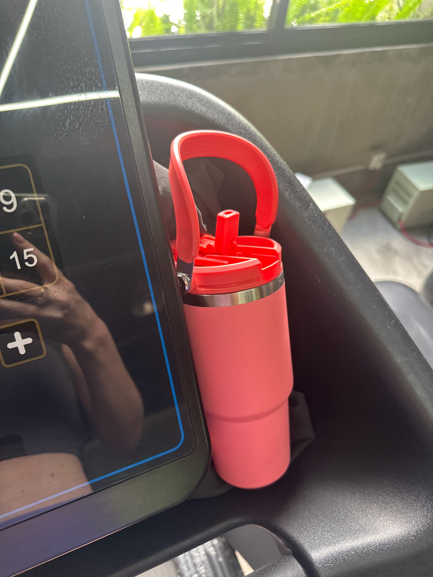 In Lalitpur, Nepal, a bright pink tumbler with a red lid and handle rests in a black cup holder attached to the side of a dark gray treadmill. The treadmill's black console is partially visible on the left, displaying white numbers and symbols against a dark background, including the number "15" and a plus sign within a square. A reflection on the console shows a person's arm and hand wearing a ring. The background features a blurred view of lush green foliage and a window, suggesting an indoor setting with an outdoor view. The treadmill is positioned on a textured gray floor.