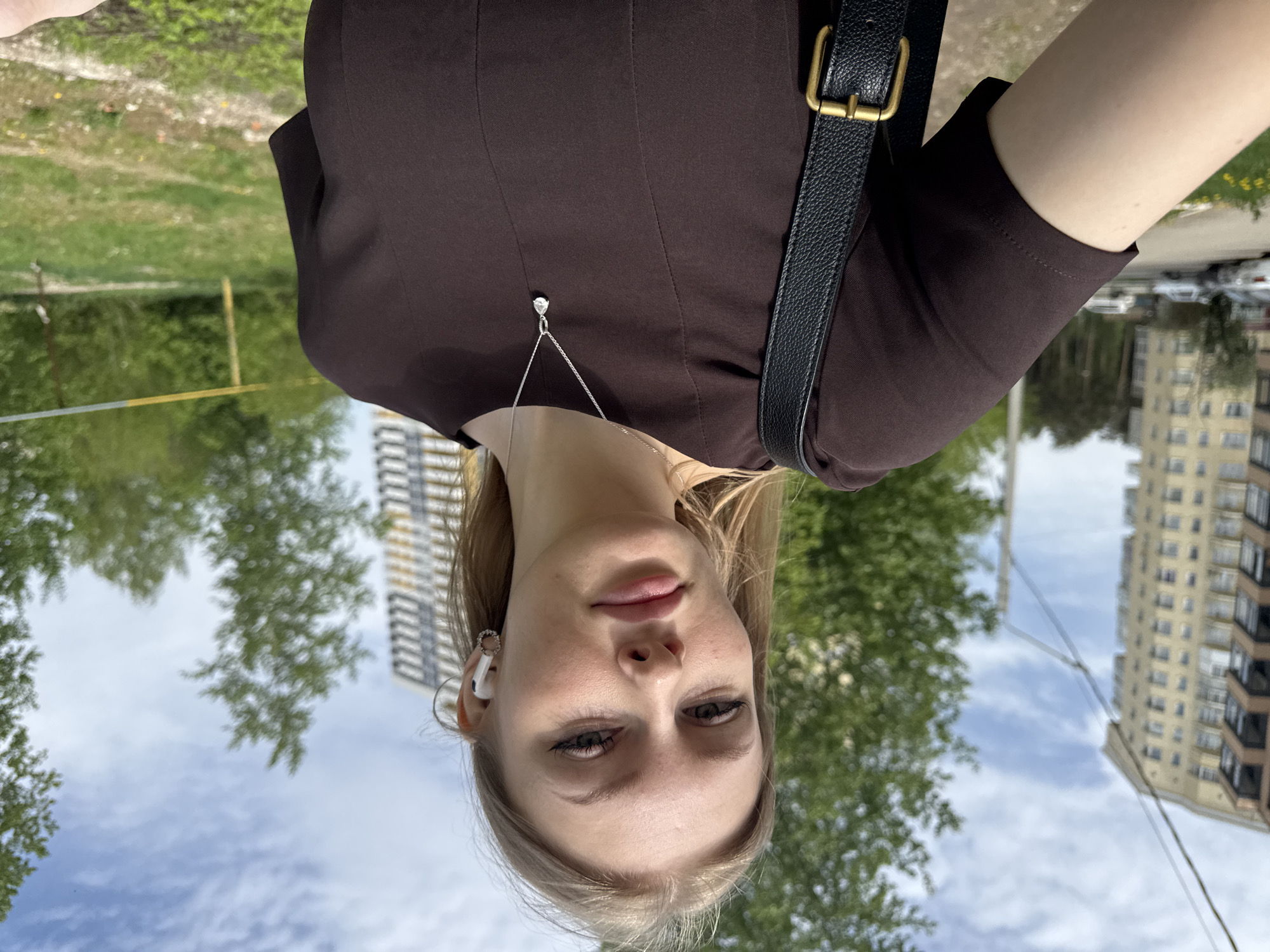 A selfie taken by a fair-skinned young woman with blonde hair, wearing a brown top and a silver necklace with a clear pendant, and an AirPod in her left ear. She is wearing a black leather shoulder bag with a gold buckle. The background is an outdoor scene in Perm, Russia, featuring buildings, trees, a partly cloudy sky, and a grassy area. The buildings suggest an urban environment.