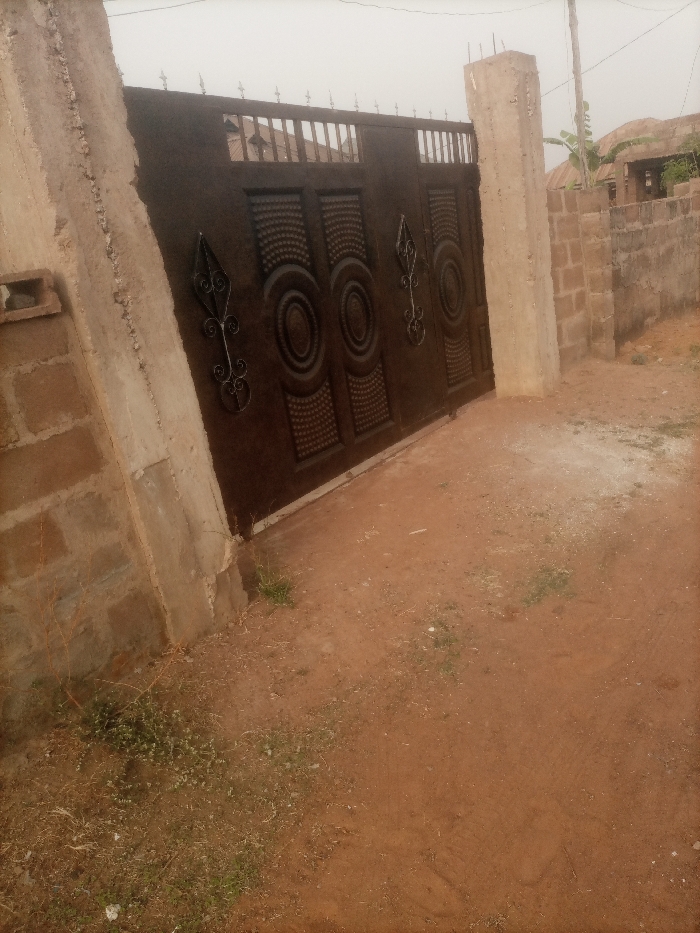 A brown metal gate, adorned with circular and spear-shaped ornaments, stands between two cement pillars. The left pillar is partially integrated with a brick wall. A brick fence runs along the right side of the gate. The ground in front of the gate is dirt, sparsely covered with small patches of grass and some dry leaves. A power line and a hint of a building are visible in the background, adding depth to the scene.