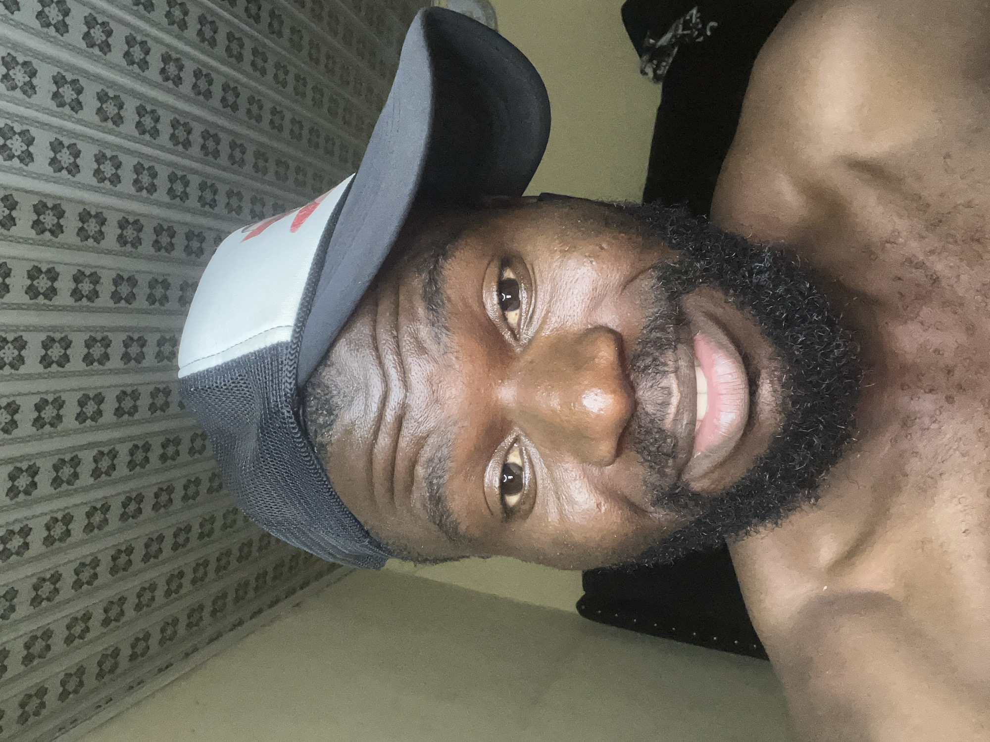 A close-up selfie shows a man with dark skin, a beard, and a trucker hat, with his upper chest visible. The background features a textured wall. The location is indeterminate without additional contextual clues.