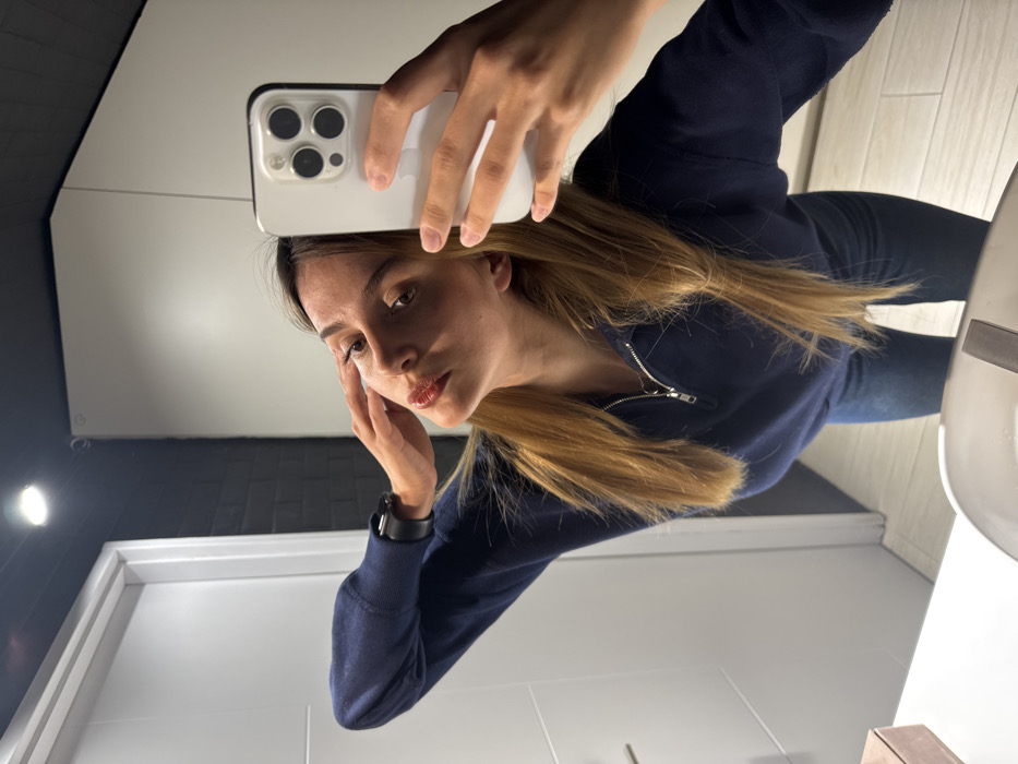 A young adult female is depicted in a mirror selfie within what appears to be a modern bathroom or washroom. The specific location within the city of Tbilisi, Georgia, cannot be determined from the image. The female has long light brown hair and wears a navy blue quarter-zip pullover and dark blue jeans. A black watch is visible on her left wrist. She holds a white iPhone, featuring three camera lenses, in her right hand, capturing her reflection. Her left hand is gently positioned with fingers touching her left temple. The environment includes a white counter with a visible portion of a sink basin in the foreground, a white door with a silver handle on the left, and a contrasting dark grey/black brick-patterned wall. The ceiling is dark and features an illuminated recessed spotlight. A small, light-colored rectangular box rests on the counter to the left of the sink.