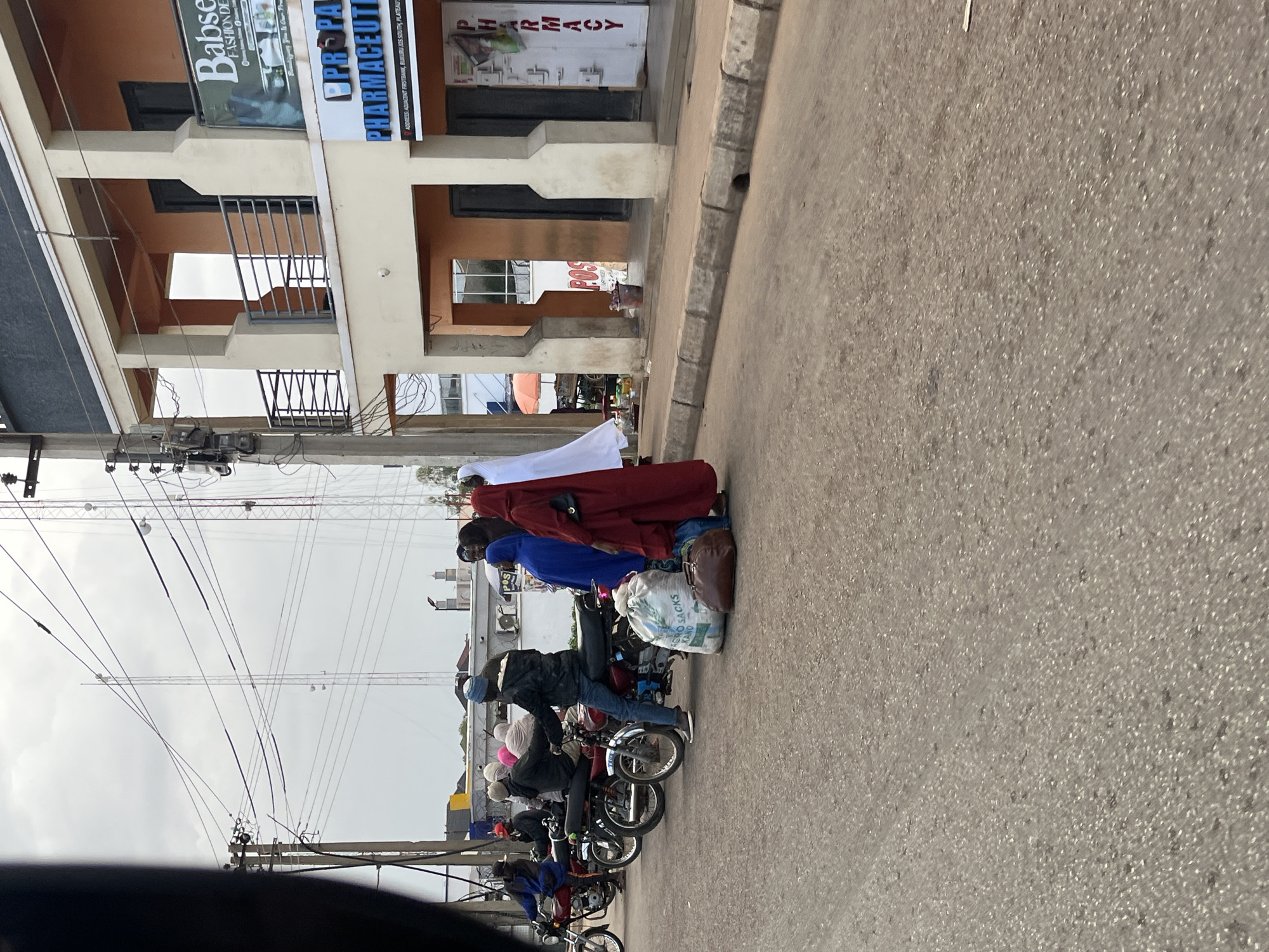 In Bukuru, Nigeria, on a cloudy day, motorcycles are parked on the right side of a paved road. Several people are gathered around the motorcycles, some seated and others standing. A white veiled figure stands next to a person in a red garment holding a dark bag. A large white sack labeled "AGRO SACKS KANO" and a brown leather bag are placed on the ground near them. In the background, a multi-story building with balconies and open doorways has signage for "Babse FASHION DEPT" and "PHARMACEUTIC PHARMACY". Electrical poles and transmission towers are visible against the overcast sky. The environment appears to be a commercial area with traffic and pedestrian activity.