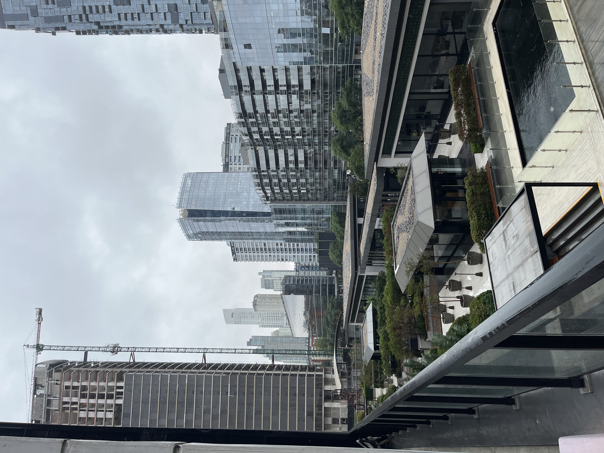 The image shows a view of Puerto Madero, Buenos Aires, Argentina. Several modern high-rise buildings are visible. The foreground features a commercial area with plantings, walkways, and what appears to be a decorative water feature. A building under construction with a crane stands on the left. The sky is overcast. There are no people visible. The image appears to have been taken from an elevated position, possibly a balcony or rooftop.