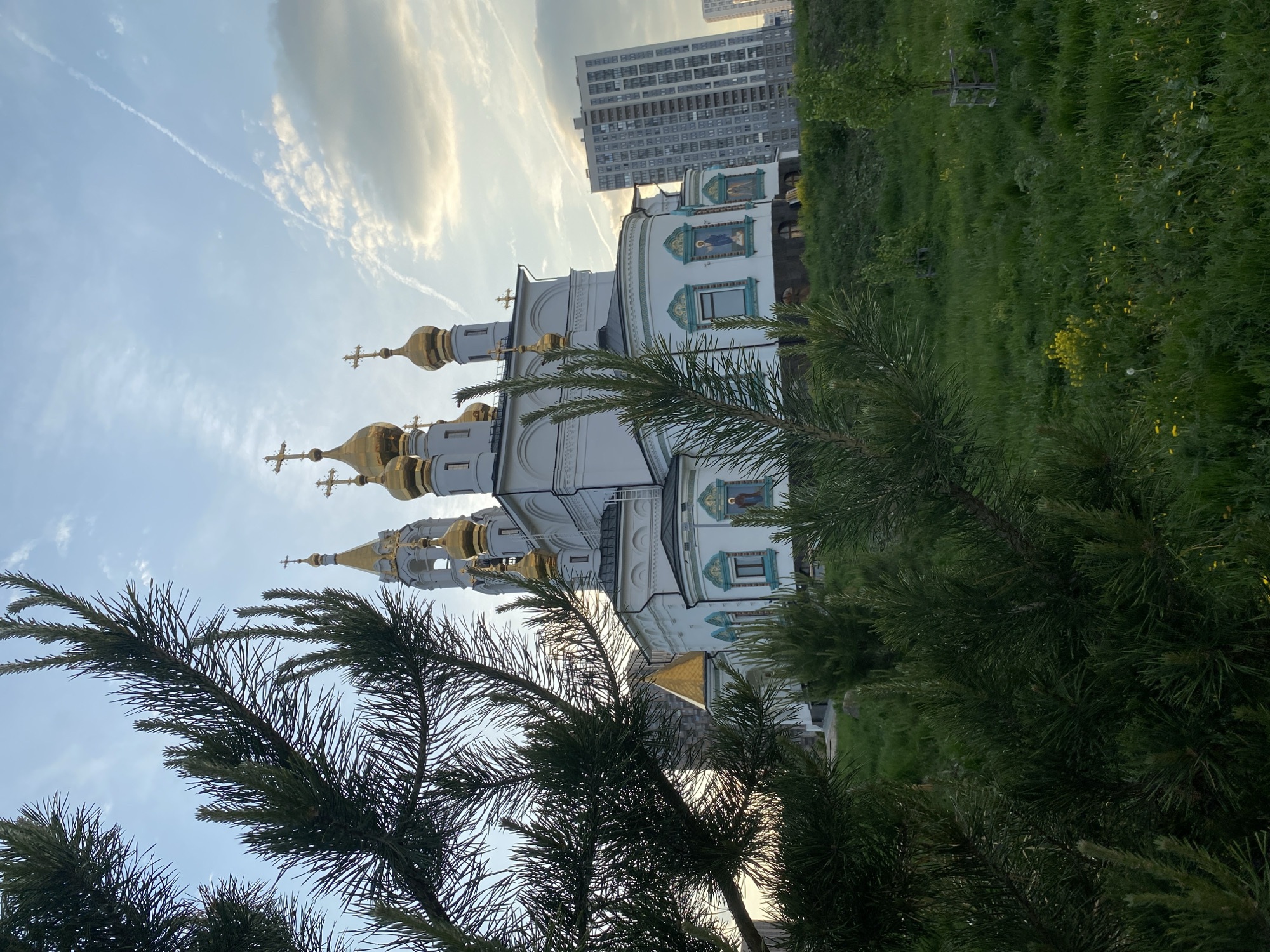 The image shows a Russian Orthodox church with golden domes and crosses in Yekaterinburg, Russia. The church is white with green and blue accents. A modern apartment building is visible in the background. Evergreen trees are in the foreground, partially obscuring the view of the church. The sky is blue with streaks of clouds. The scene appears to be taken during daytime.