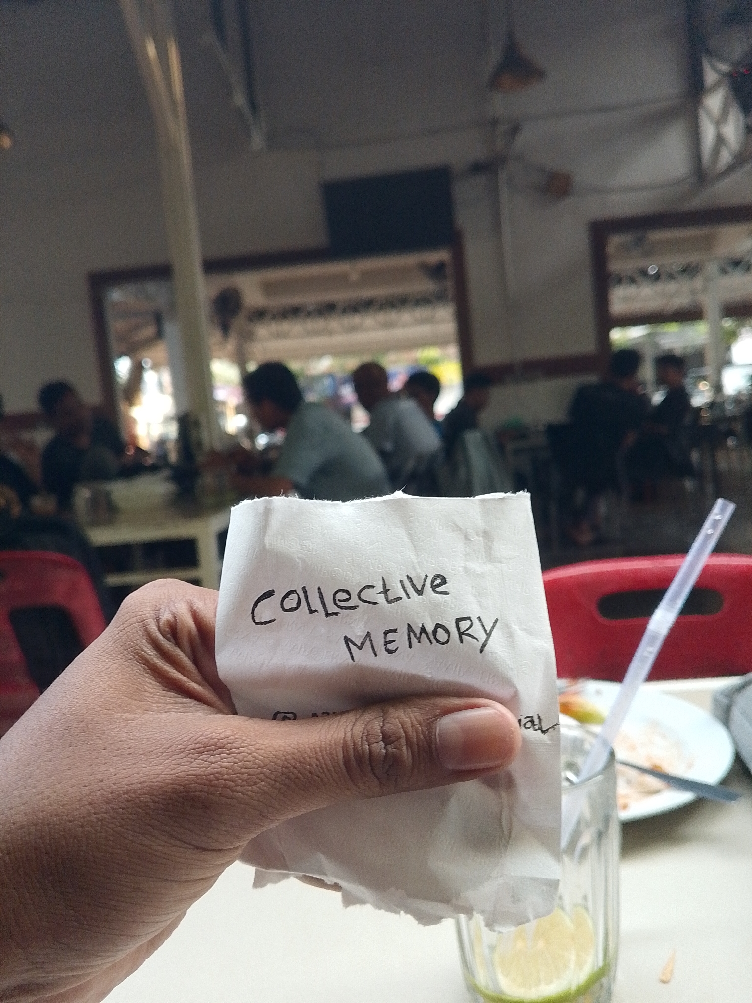 A close-up, medium shot in what appears to be a cafe or restaurant in Lhokseumawe, Indonesia. The focus is on a white napkin held in a brown-skinned hand. "Collective MEMORY" is handwritten in black ink on the napkin. Beside the hand is a glass with a sliced lime inside and a white plastic straw. There is a white plate with food remnants nearby and a table with a red chair. In the background, a casual, blurred scene shows several people sitting at tables, suggesting a social setting within a typical indoor public space. Windows and decor visible in the background give the impression of a relaxed, local establishment. A television is visible in the background.