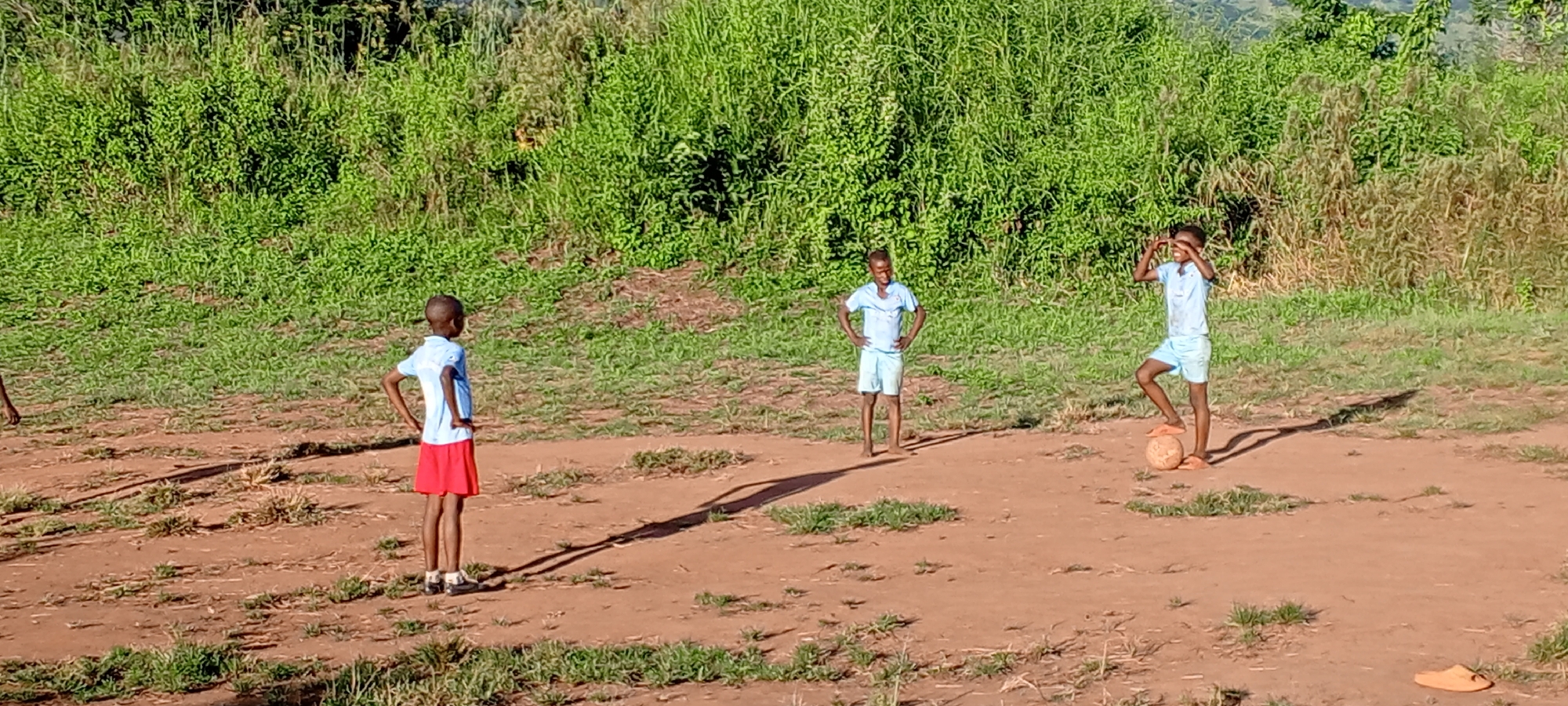 Three boys stand on a dirt ground in Ngalula, Democratic Republic of the Congo. The boy on the left wears a light blue shirt and red shorts. The middle boy wears a matching light blue shirt and shorts and has his hands on his hips. The boy on the right also wears a matching light blue shirt and shorts. He is standing with one foot on a soccer ball, and his hands are above his head. Overgrown green bushes surround the field.