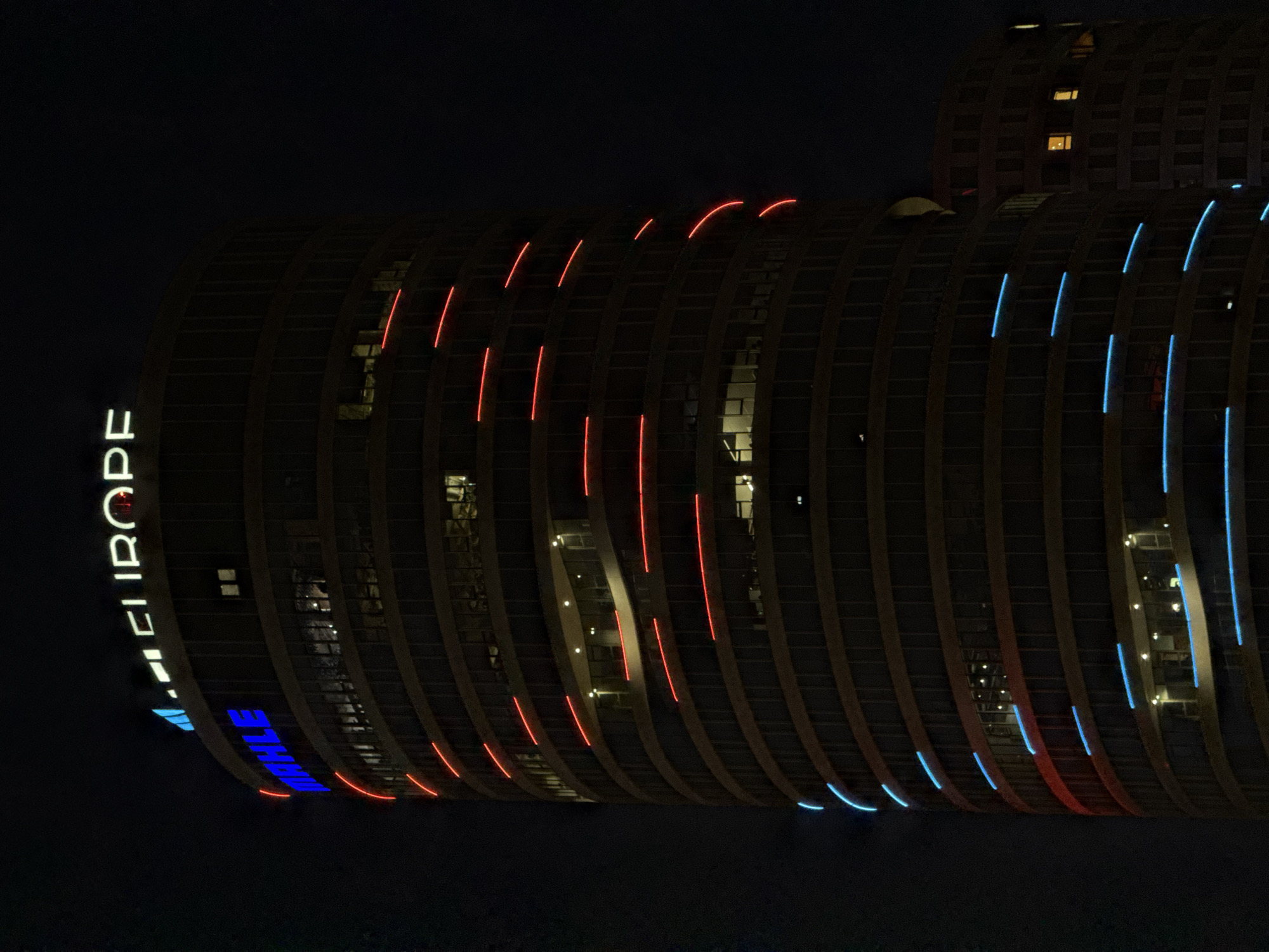 The image displays a modern, tall building in Belgrade, Serbia, illuminated at night. The structure features a distinctive curved facade, characterized by a series of vertical, rib-like architectural elements. The deep night sky forms a dark backdrop, causing the building's lights to stand out vividly.
The facade is adorned with dynamic LED lighting, featuring alternating segments of vibrant red and cool blue light strips that trace the building's curves. These lights are mostly vertical or follow the arc of the architectural elements, creating a striking visual pattern across the building's dark surface. Scattered windows are also lit from within, suggesting activity inside.
Prominently displayed vertically along the left side of the building, the word "EUROPE" is illuminated in bright white capital letters, reading from bottom to top. Below this, partially visible and illuminated in blue, is the text "MAHLE". The building appears to be a sleek and contemporary urban landmark, possibly an office tower or hotel.
The time of day is clearly night, with no visible clouds, indicating clear weather. The calm and still scene emphasizes the architectural design and artificial illumination, contributing to a modern and eye-catching aesthetic.