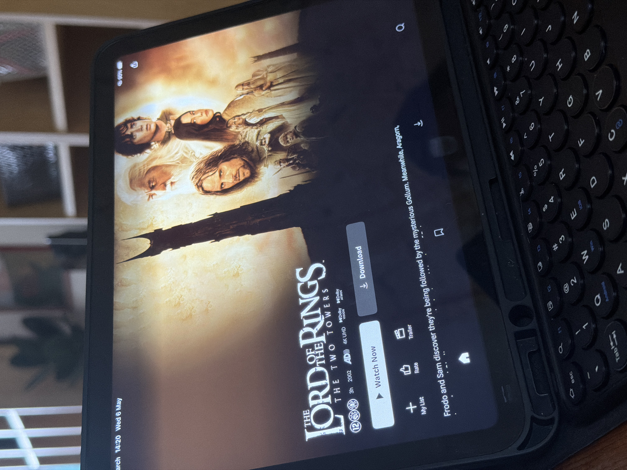 Here is a matter-of-fact description of the scene in the image:
The image shows a tablet displaying the Netflix menu for the movie "The Lord of the Rings: The Two Towers". The screen shows the film's cover art, alongside the "Watch Now" button and other options like "Download" and "Trailer". The timestamp on the tablet indicates it is Wednesday, May 6th, at 14:20. Part of a black keyboard is attached to the tablet, covering much of the right side of the screen. A blurred bookcase is visible in the background. The location is not indicated in the image.