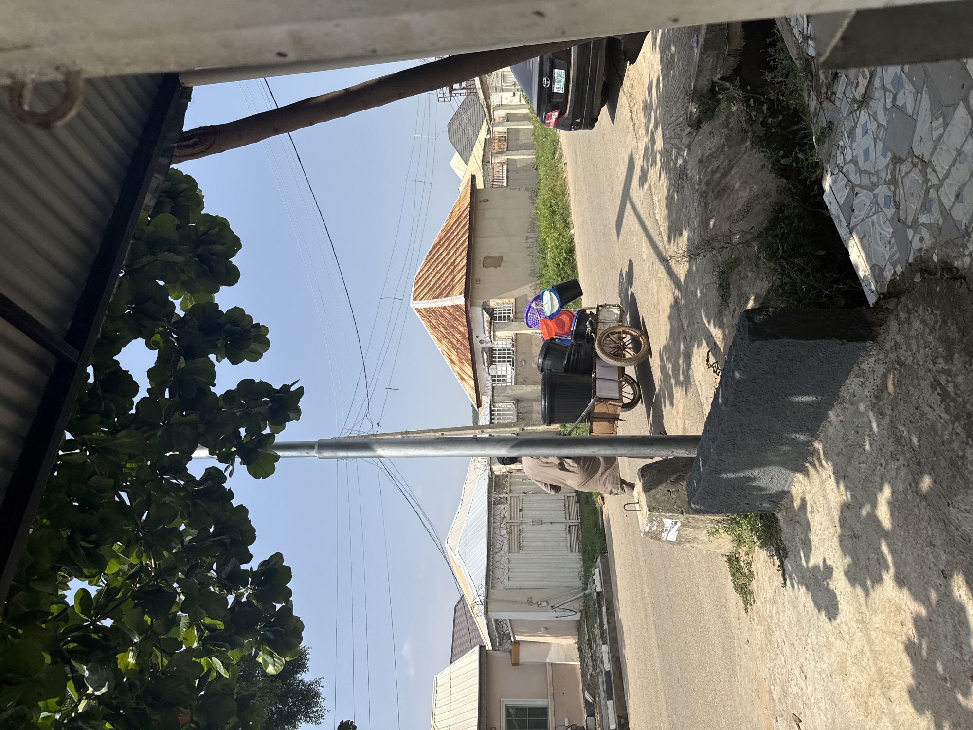 This media file captures a street scene in Abuja, Nigeria, under a clear blue sky, suggesting a bright, sunny day. The image is oriented vertically, with a portion of a corrugated metal roof and a tree with large, dark green leaves dominating the left foreground. A utility pole with multiple wires runs vertically through the frame, casting shadows on the ground.
In the middle ground, a cart is laden with various colored plastic buckets, some appearing new and stacked neatly, others possibly used. The cart itself has wooden components and a spoked wheel. A person, clad in a light brown, loose-fitting garment, is partially visible behind the cart, suggesting they are either pushing or attending to it.
The background reveals a street lined with buildings. One building has a distinctive rusted, corrugated metal roof. Another building features a white gate with barbed wire on top, indicating a measure of security. A black car with a visible license plate, GNF104ET, is parked further down the street. The presence of power lines and the style of the houses suggest a residential or semi-commercial area within Abuja. The overall impression is of a typical day in a Nigerian urban environment, with daily activities, such as the movement of goods via cart, being a visible element.