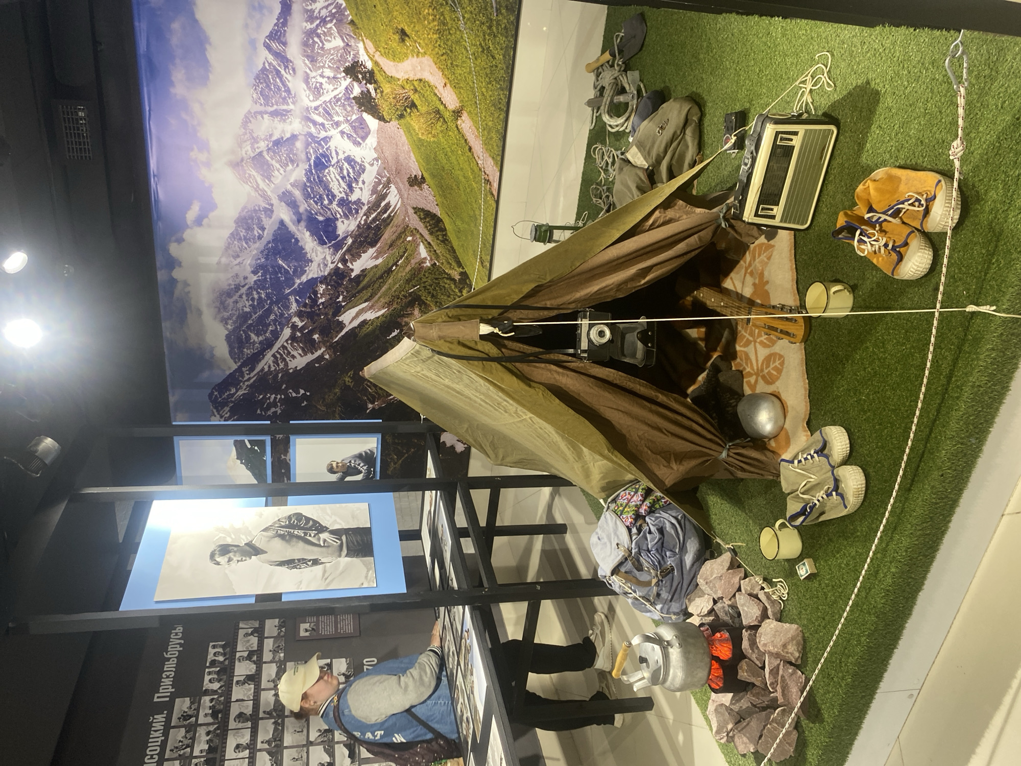 This image depicts a museum exhibit in Yekaterinburg, Russia, likely at the Yeltsin Center, showcasing elements of mountaineering and outdoor adventure. In the foreground, a green artificial turf surface simulates a campsite. A brown and green tent is pitched, with a vintage camera hanging inside. Various camping gear is arranged around the tent, including a kettle on a simulated campfire with red coals, a backpack, a lantern, ropes, a shovel, a radio, a mug, and a pair of hiking boots. Behind the campsite, a large mural displays a majestic mountain landscape with snow-capped peaks and a winding path. To the left of the campsite, a display table exhibits framed photographs and documents related to mountaineering history. A woman wearing a cap and a denim jacket is observing these exhibits. The overall scene is designed to evoke a sense of historical exploration and the spirit of adventure associated with mountainous regions.