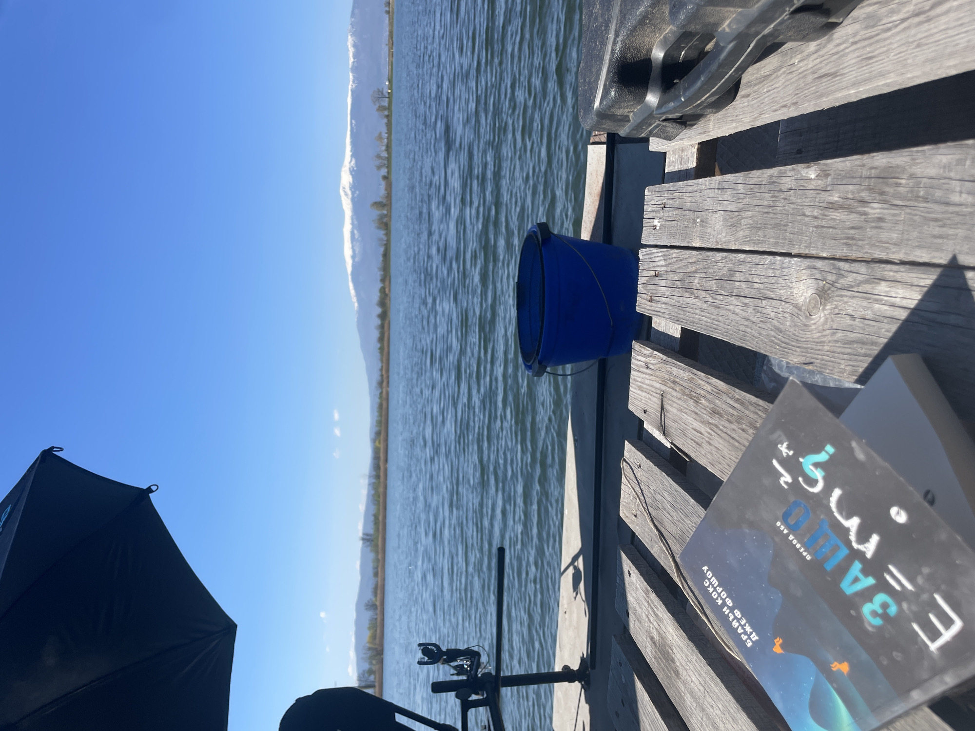 The image shows a view from a wooden dock or platform overlooking a body of water with mountains in the background under a clear, blue sky.
On the dock, there is a blue bucket, a fishing rod stand, and a book with text and imagery on its cover. The text is in Bulgarian and reads "Е = mc2 ЗАЩО Е ТОЛКОВА ВАЖНО?", which translates to "E=mc2 WHY DOES IT MATTER?". The authors are listed as Brian Cox and Jeff Forshaw. Part of a black umbrella is also visible in the lower left corner of the frame.
The location is not explicitly identifiable as Sofia, Bulgaria from the image alone. There is no visual evidence to confirm the location.