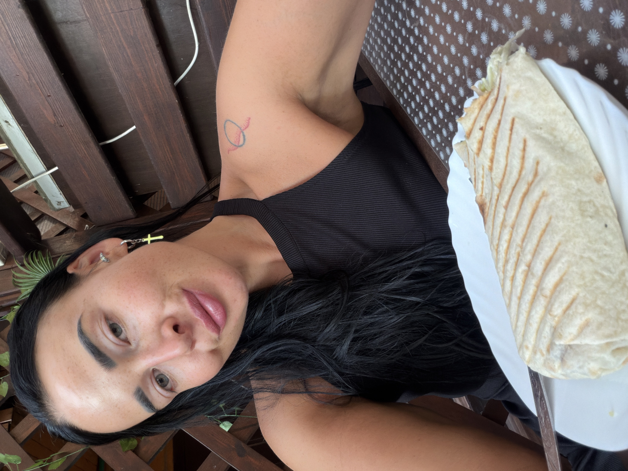 This is a close-up, vertically oriented, selfie-style photograph of a woman with long, dark hair and light-colored eyes. She is wearing a black sleeveless top and a cross earring in her left ear. A small, abstract tattoo in blue and red is visible on her upper right arm. She has a light dusting of freckles across her nose and cheeks.
In the foreground, on the right side of the image, is a white plate with a shawarma or wrap on it. The wrap is golden brown with grill marks. The background features wooden slats, suggesting an outdoor setting, possibly a patio or deck. A dark brown tablecloth with a white pattern is visible behind the plate.
The lighting suggests it is daytime. The woman's expression is neutral, with her lips slightly parted. The overall scene is casual and appears to be a meal taken outdoors. No text is visible in the image.