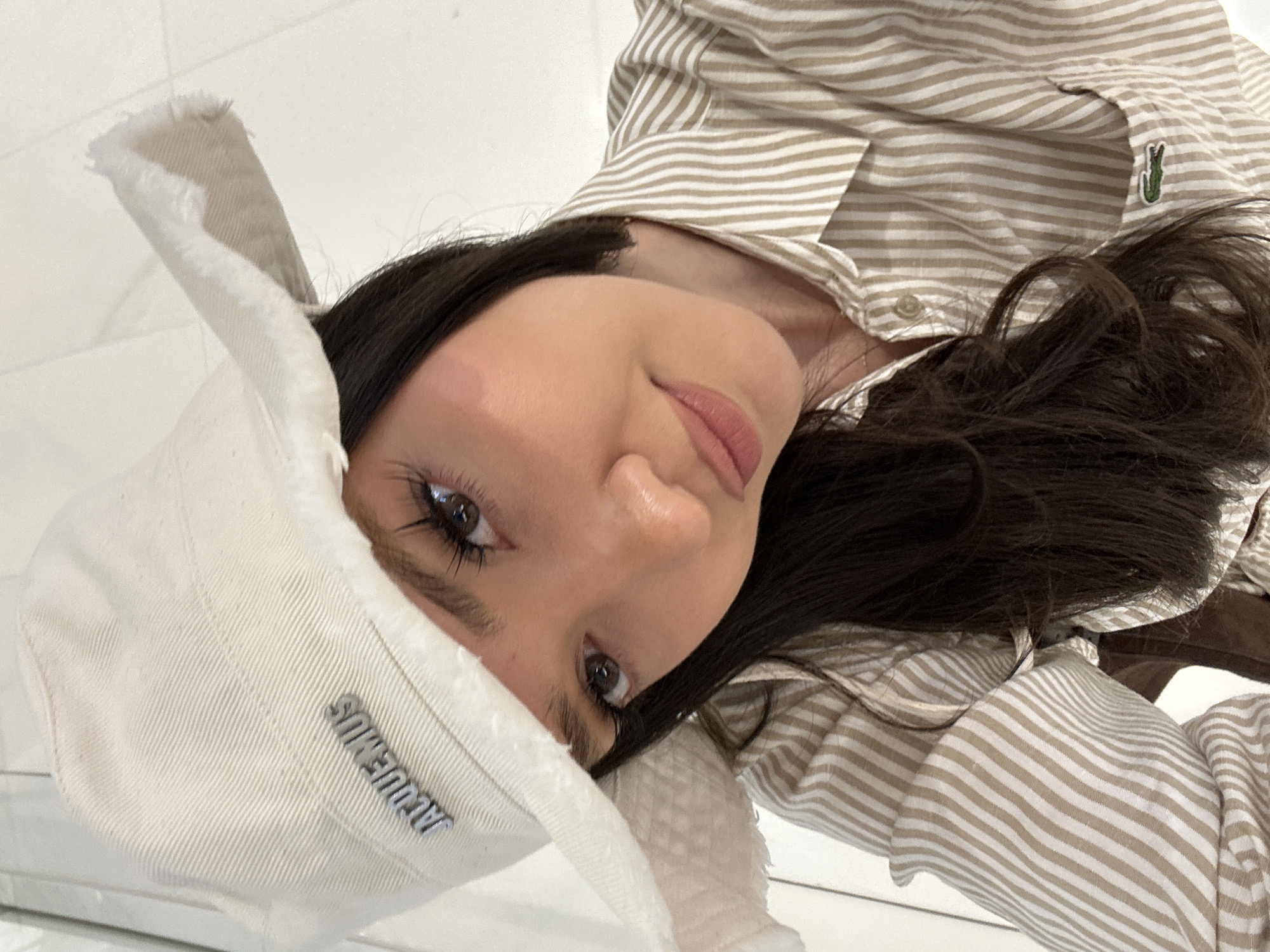 The image, oriented sideways, is a close-up portrait of a young woman with long, dark hair. She looks directly at the camera with a calm, soft expression, featuring dark eyelashes and nude or natural-toned lipstick. She is wearing an off-white or cream-colored bucket hat with a distressed or frayed brim. The brand name "JACQUEMUS" is clearly visible in silver metallic letters on the front of the hat. Her attire also includes a collared button-down shirt with light beige and white horizontal stripes, featuring the distinctive green crocodile logo of Lacoste on the left side. The background is a plain, bright, light-colored surface, suggesting an indoor setting with clean, minimalist aesthetics. The photograph appears to be a selfie, capturing her from a low angle. The location context provided for this image is Dubai, United Arab Emirates.
