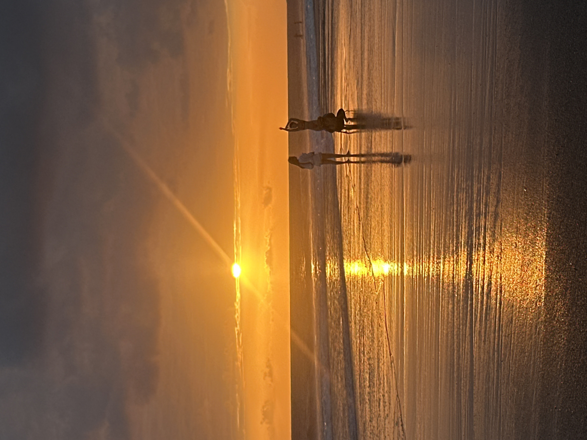 In North Kuta, Indonesia, a sunset illuminates a beach with a vibrant orange glow. The sun, partially obscured by clouds, casts a bright reflection on the wet sand. Two individuals are silhouetted against the sunset. One stands upright, facing away from the camera, while another is in a crouching position with another person standing on their shoulders, with arms raised. The ocean is visible in the background with gentle waves.