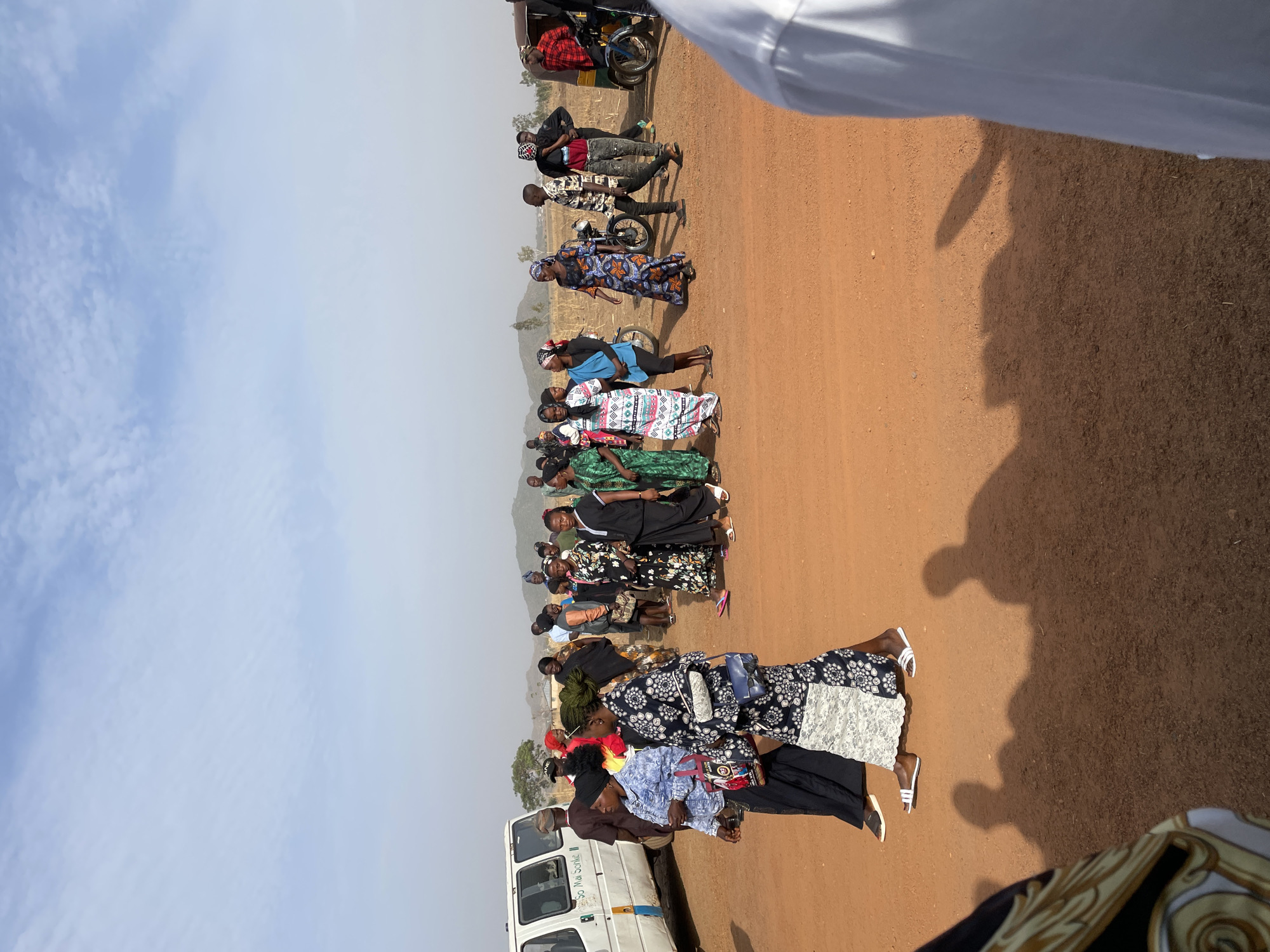 The image captures a gathering of people, primarily women, standing in a line along a wide, reddish-brown dirt road under a clear, bright sky. Distant hills or mountains are visible in the background, suggesting a rural or semi-rural setting in Du, Nigeria.
The majority of the women are dressed in colorful, patterned long dresses, while a few men are also present. A white passenger van or minibus is parked on the left side of the frame, and several motorcycles and a tricycle (keke napep) are visible further down the road, indicating the presence of transportation. The strong, distinct shadows cast by the people and objects on the ground suggest bright daylight, likely midday or early afternoon.
The people appear to be patiently waiting or queuing for an unspecified activity, possibly related to the vehicles or an event. On the side of the white van, the text "So Mai Sarke ||" is legible. The foreground shows a partial view of a white garment or fabric, and a large shadow, likely cast by the person taking the photo, is prominent on the dirt. The scene depicts an outdoor communal gathering on a sunny day.